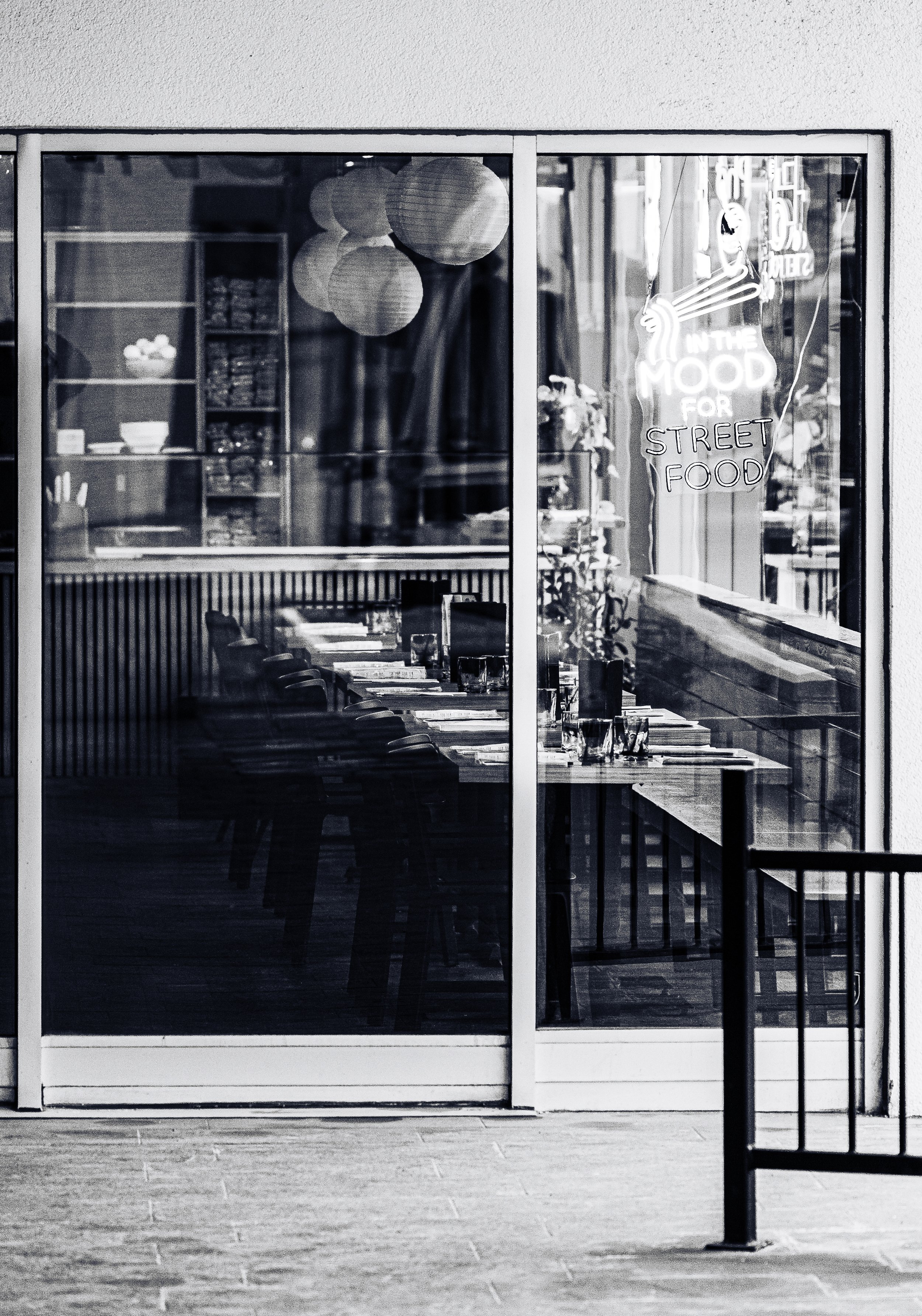 Black and white photo of a restaurant through a glass window with a neon sign that reads "In the Mood for Street Food" and paper lanterns hanging inside.