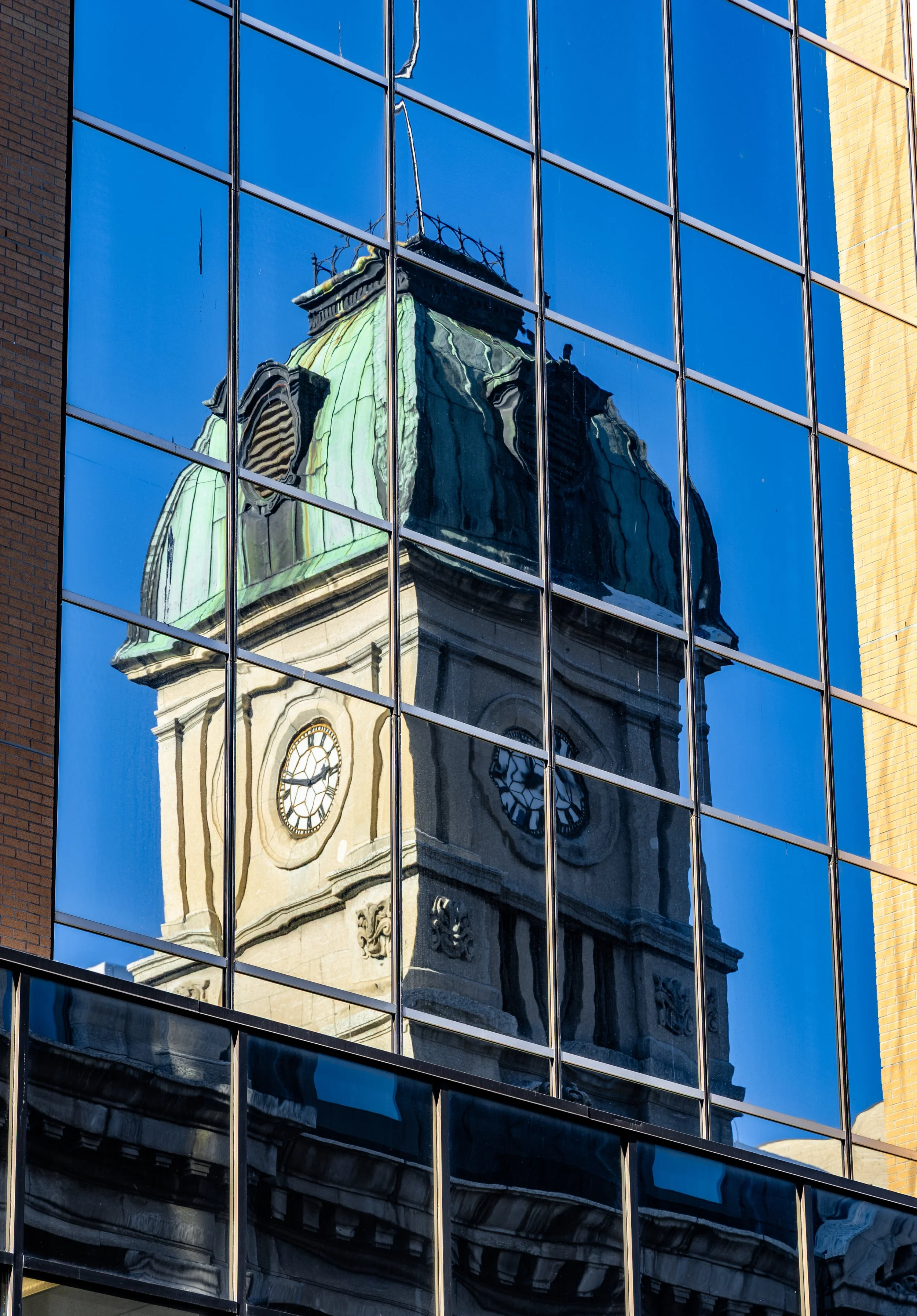 Reflection of a historic clock tower with a green dome in the glass windows of a modern building.