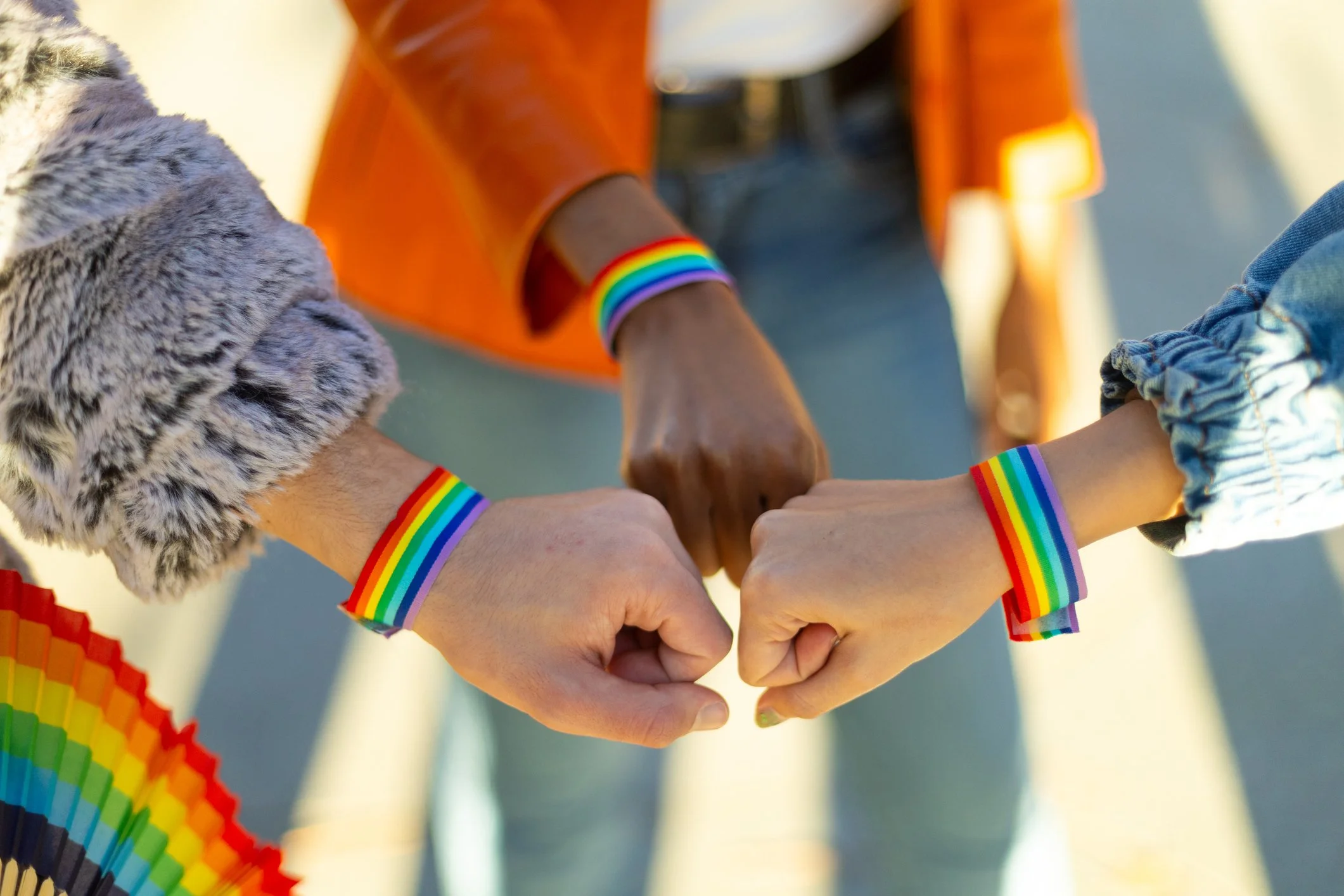 Multiple hands joined together in a circle, each wearing rainbow-colored bracelets, symbolizing diversity and unity, with rainbow-themed decorations and a person wearing a rainbow wristband.