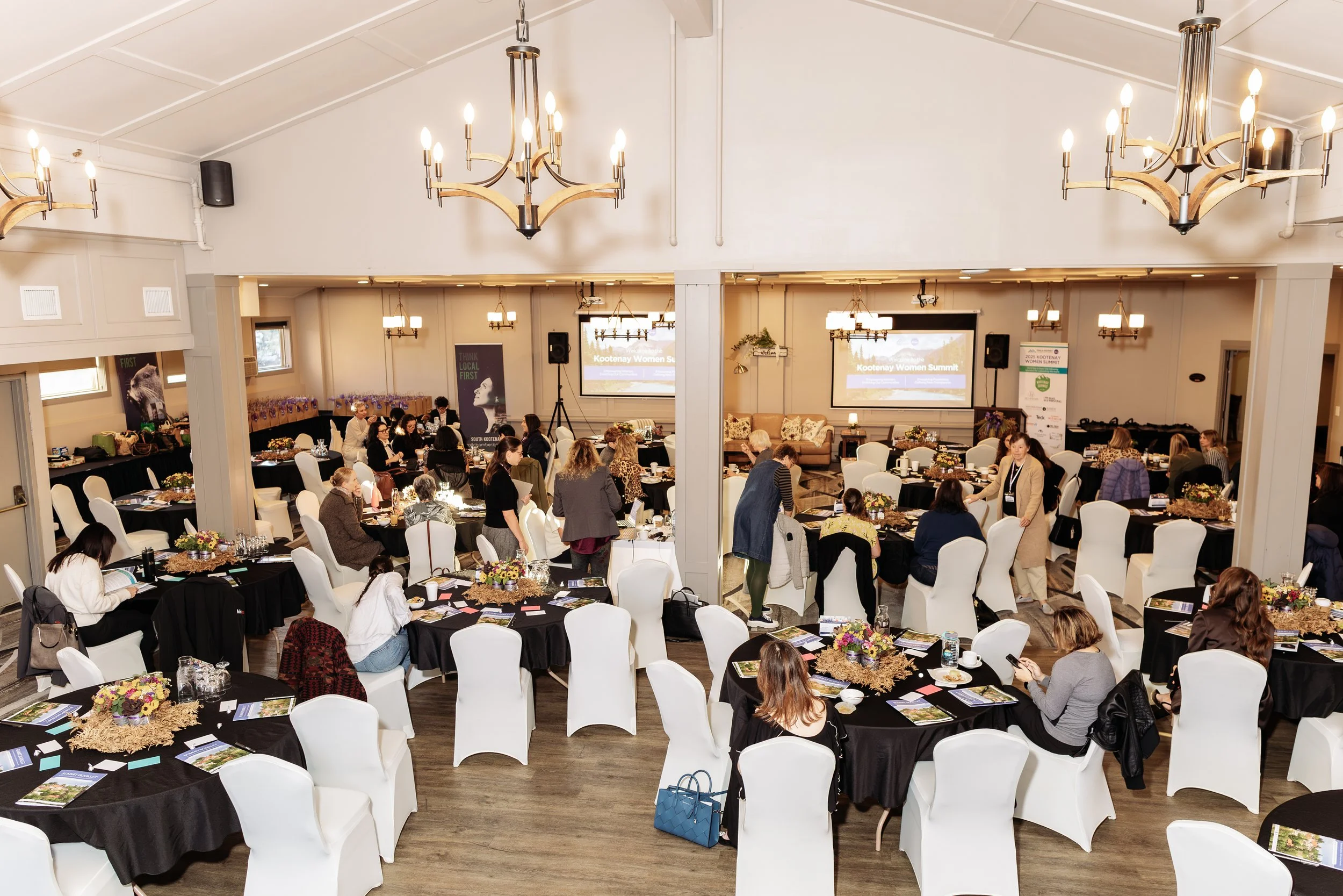 A large banquet hall filled with round tables covered with black tablecloths, each decorated with a flower centerpiece, and white chairs surrounding them. People are sitting and talking at the tables; some are standing and walking around. The front of the room has a screen with a presentation titled "Kootenay Women Summit," and there are banners and speakers on the stage. The hall has high ceilings with white walls and multiple chandeliers hanging from the ceiling.