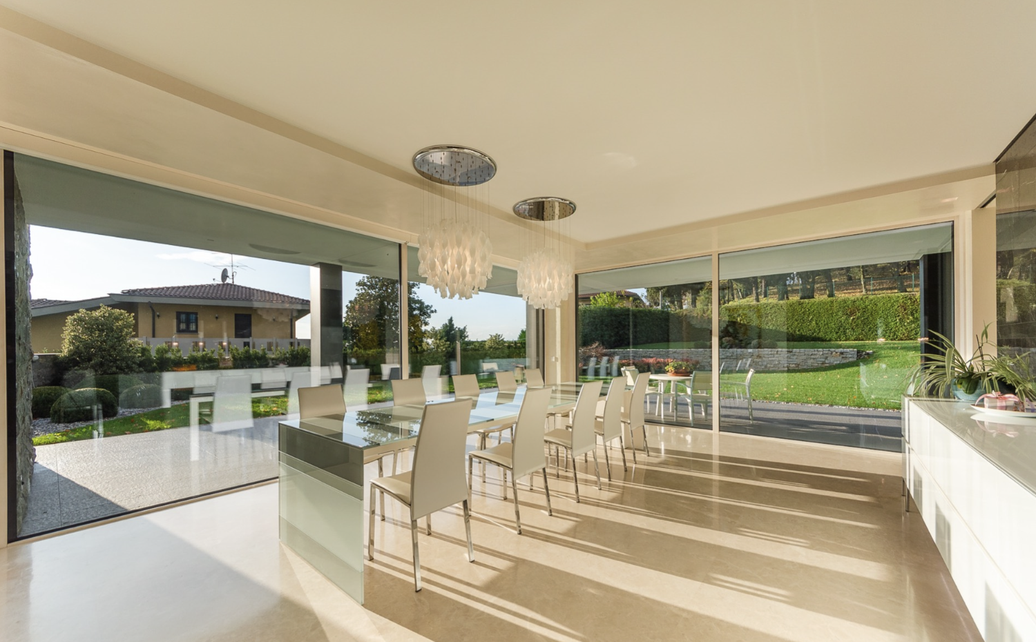 Modern dining room with glass walls, a long glass table, white chairs, and decorative light fixtures, overlooking a landscaped outdoor area.