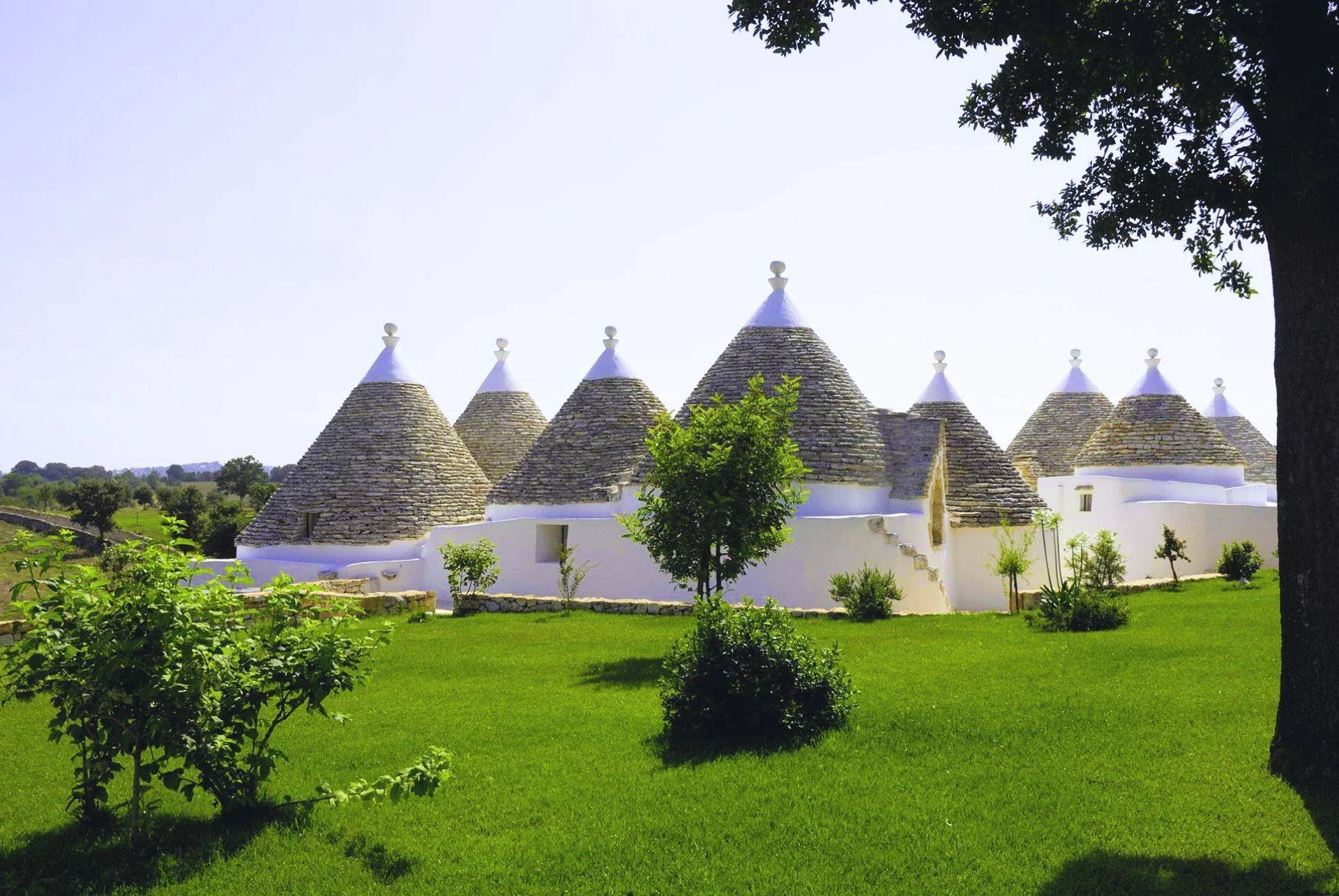 A traditional Italian Trullo with conical stone roofs and white walls, surrounded by green grass and trees.