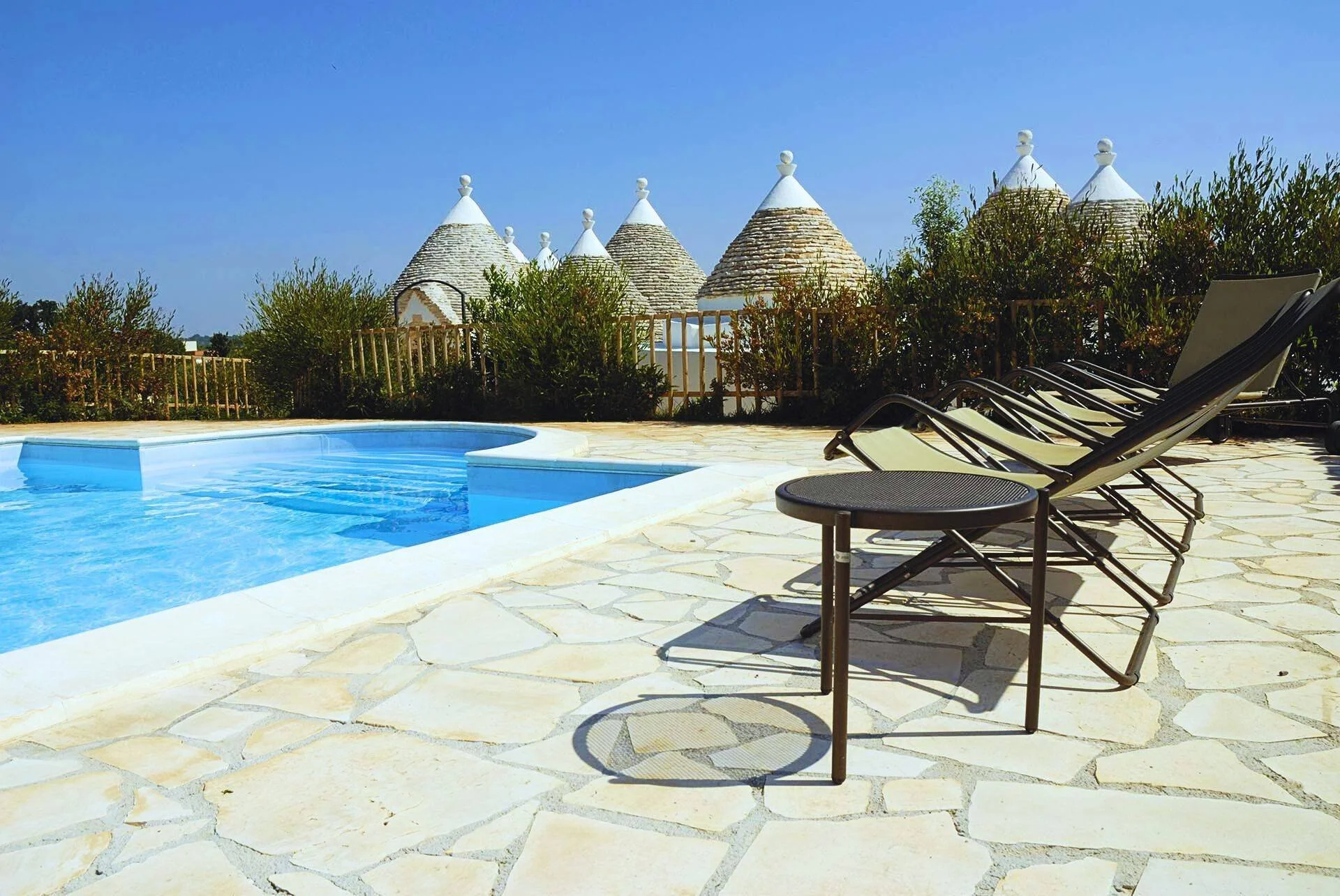 Swimming pool with four lounge chairs and a small table on a stone patio, over a fence with bushes and traditional cone-shaped buildings in the background under a clear blue sky.
