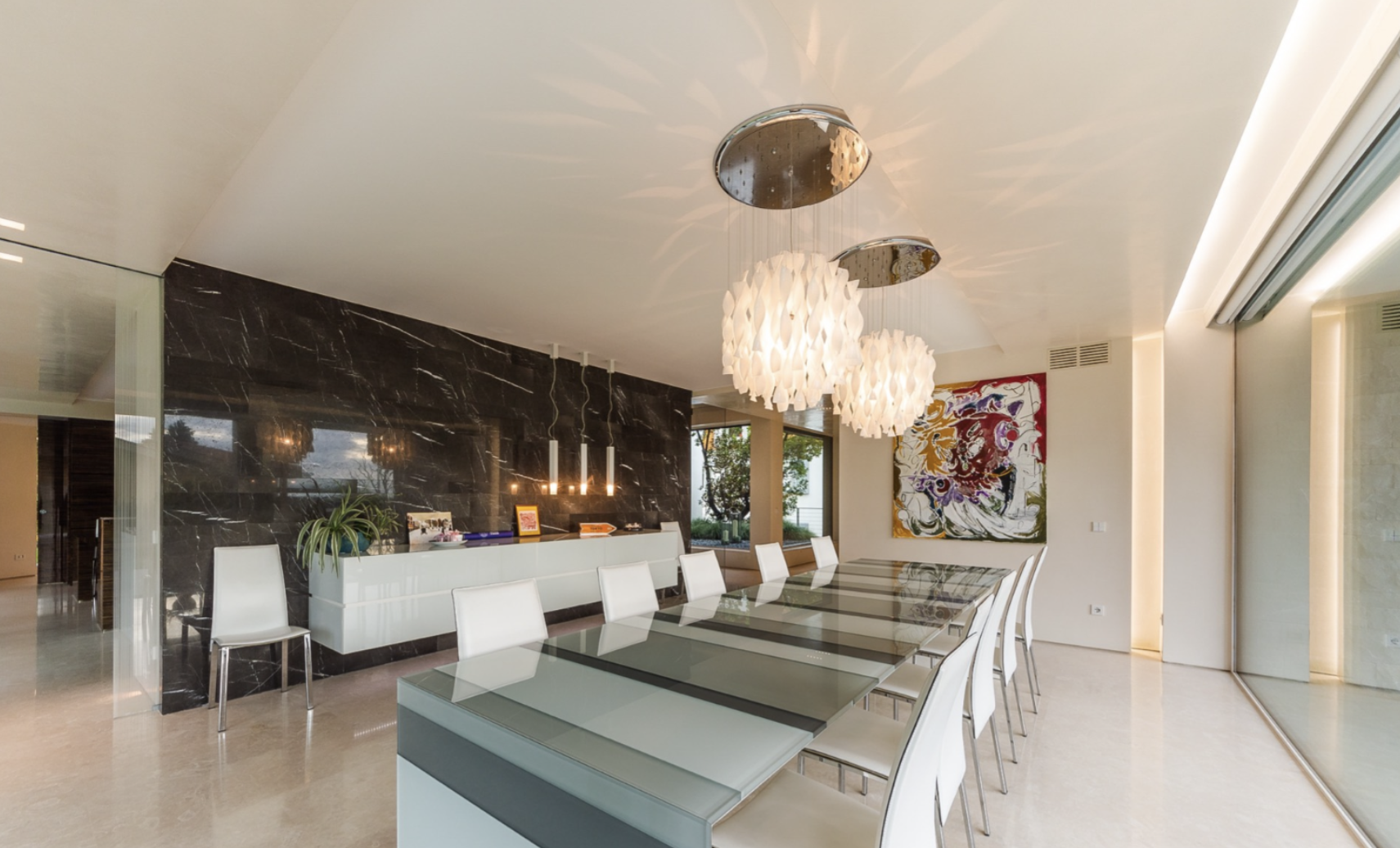 Modern dining room with glass-topped table, white chairs, abstract artwork on the wall, and decorative pendant lights.