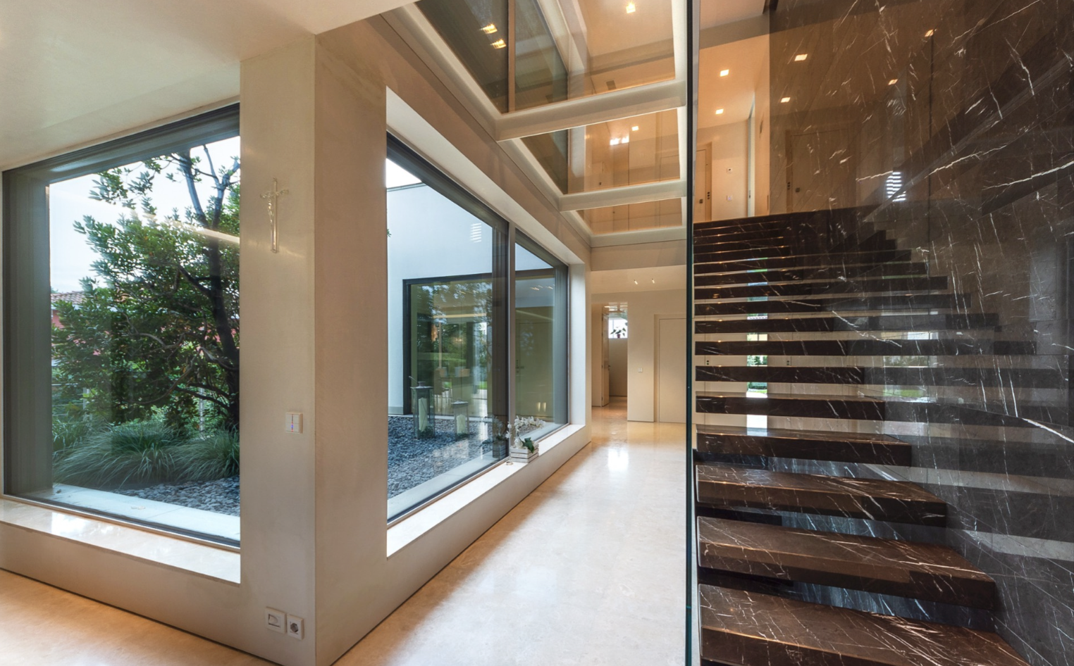 Modern interior of a house with large windows showing a garden, marble staircase, and a glass ceiling.