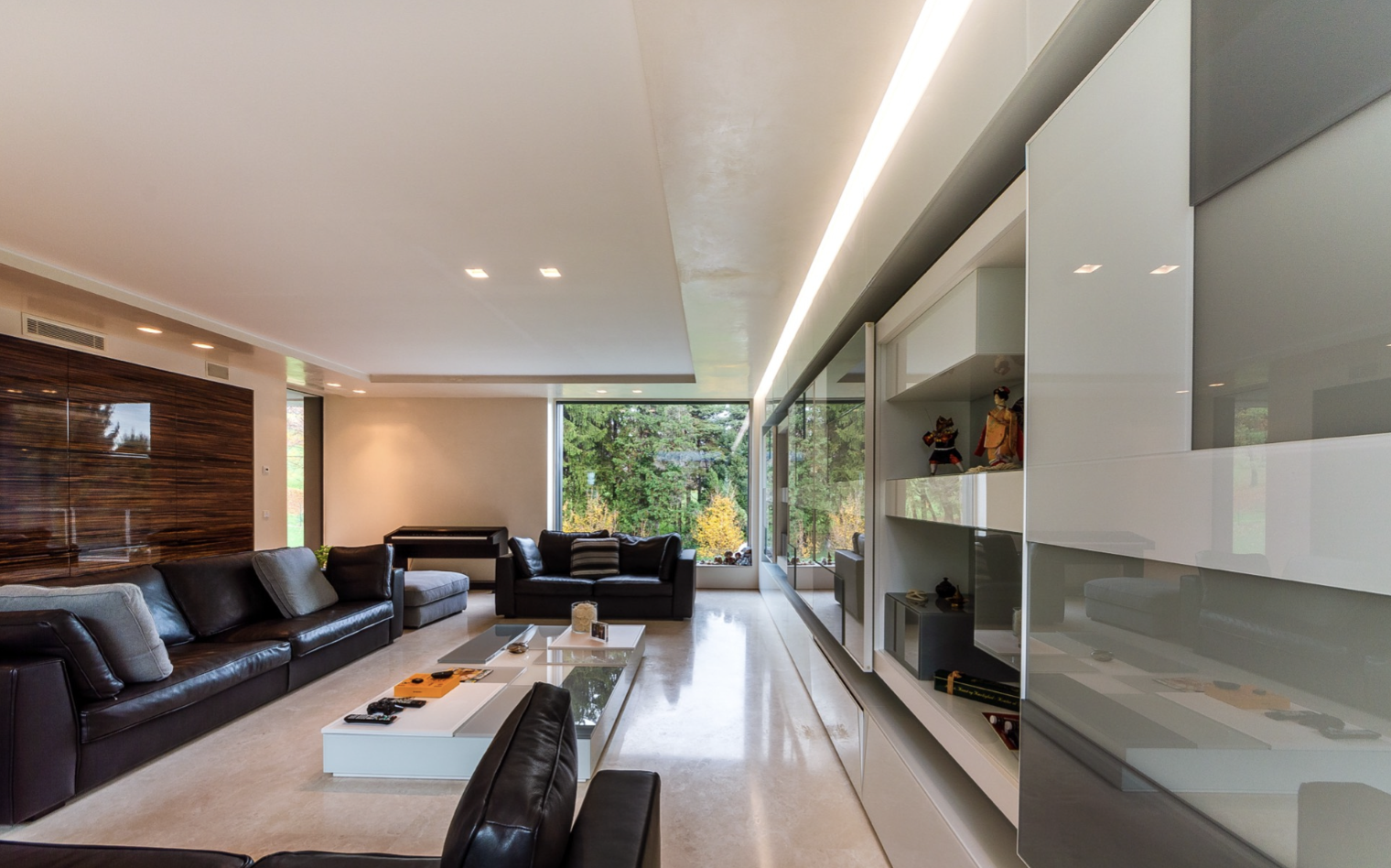 Modern living room with black leather sofas, a white coffee table, and large windows showing trees outside.