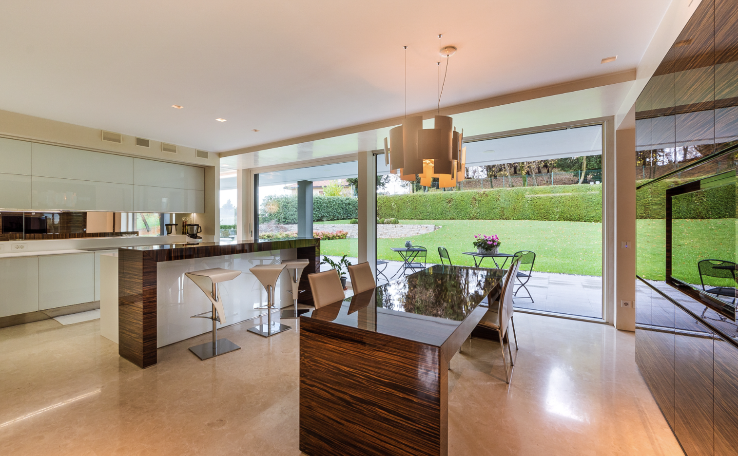 Modern kitchen and dining area with large glass sliding doors opening to a lush green backyard with patio furniture and flower pots.