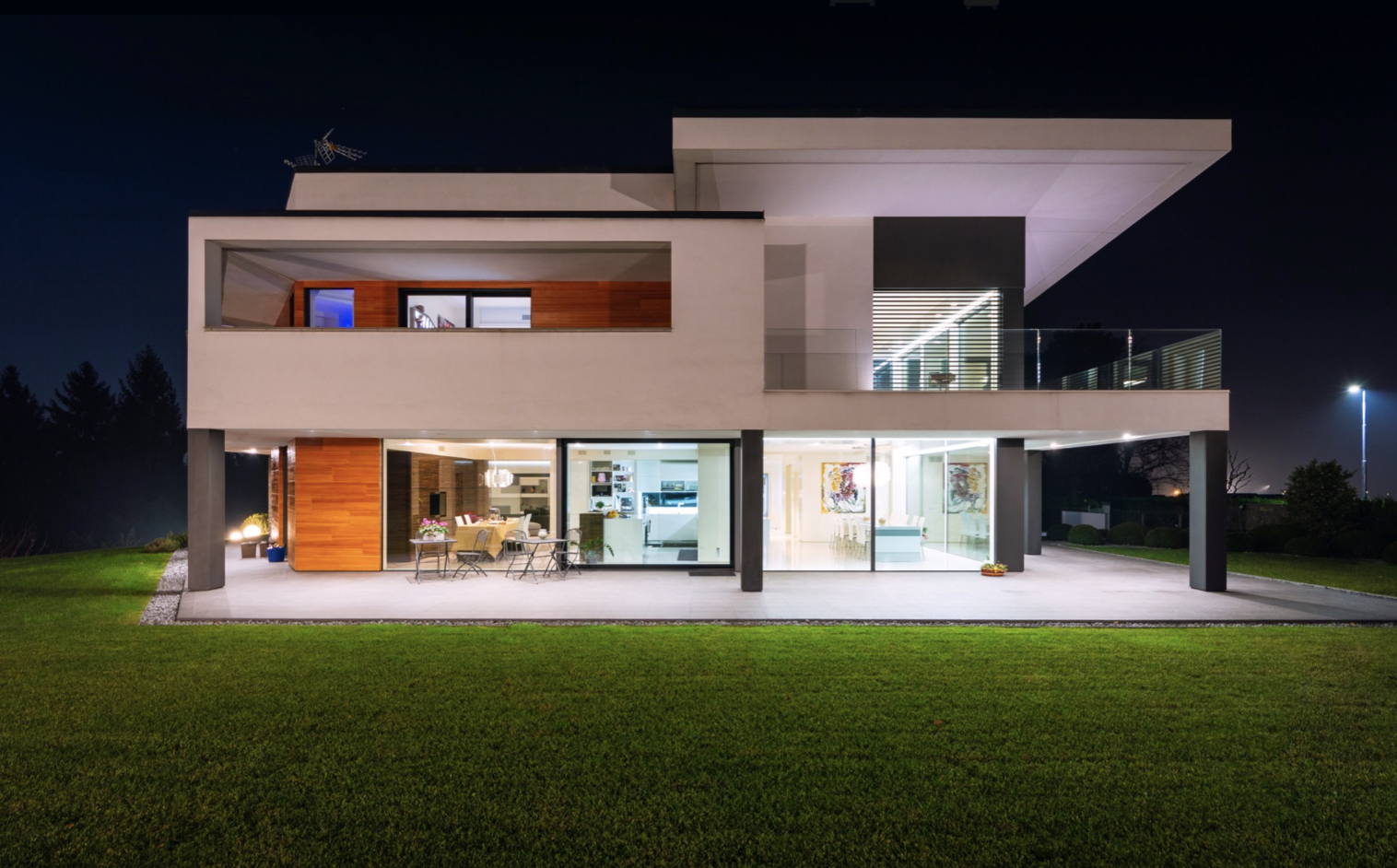Modern two-story house illuminated at night, with large glass windows and a balcony, surrounded by a well-maintained lawn.