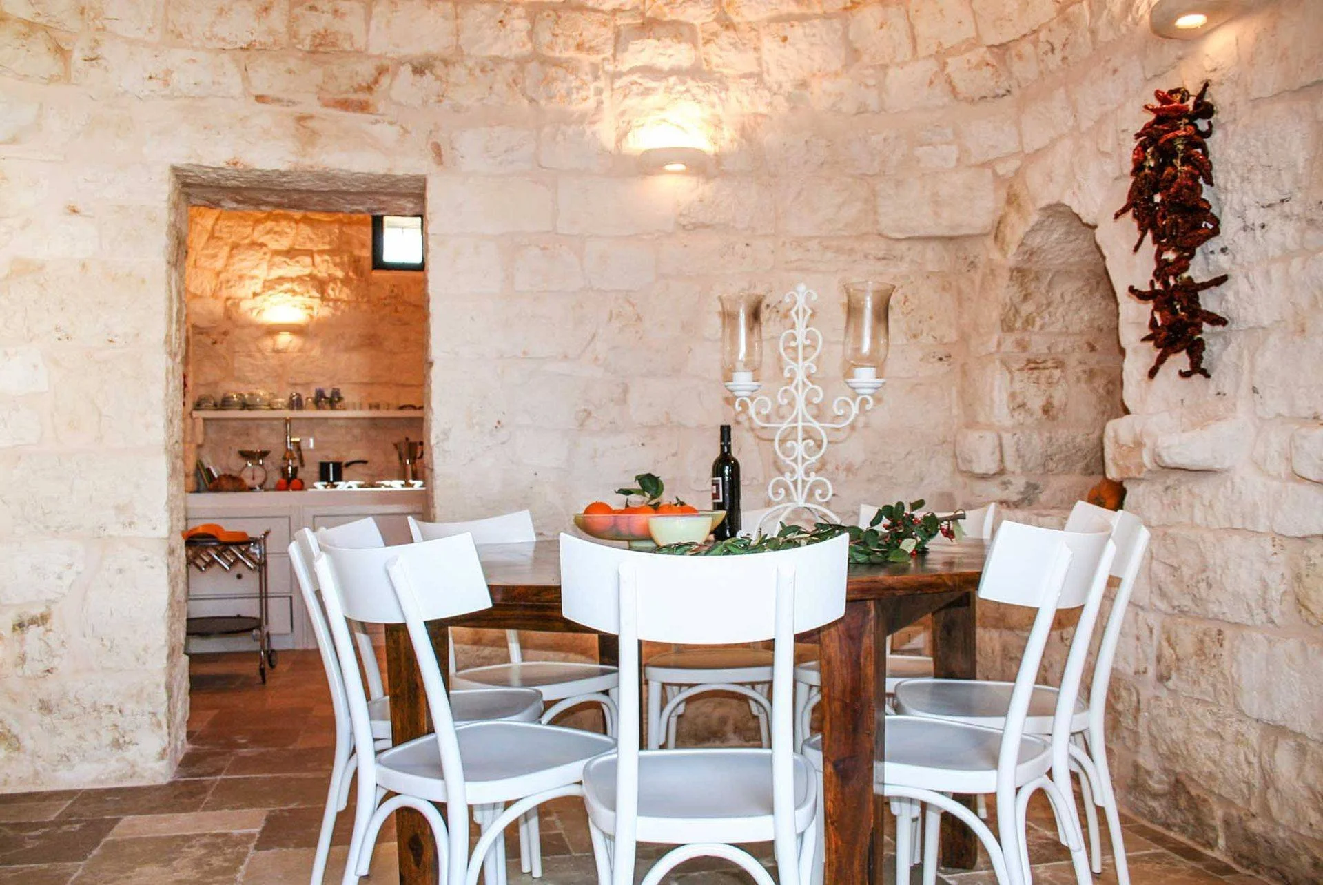 A dining room with stone walls, a round wooden table, and white chairs. The table has a bowl of fruit, a bottle of wine, and a decorative white candelabra. There is a doorway leading to a kitchen area with a window, small kitchen appliances, and a co