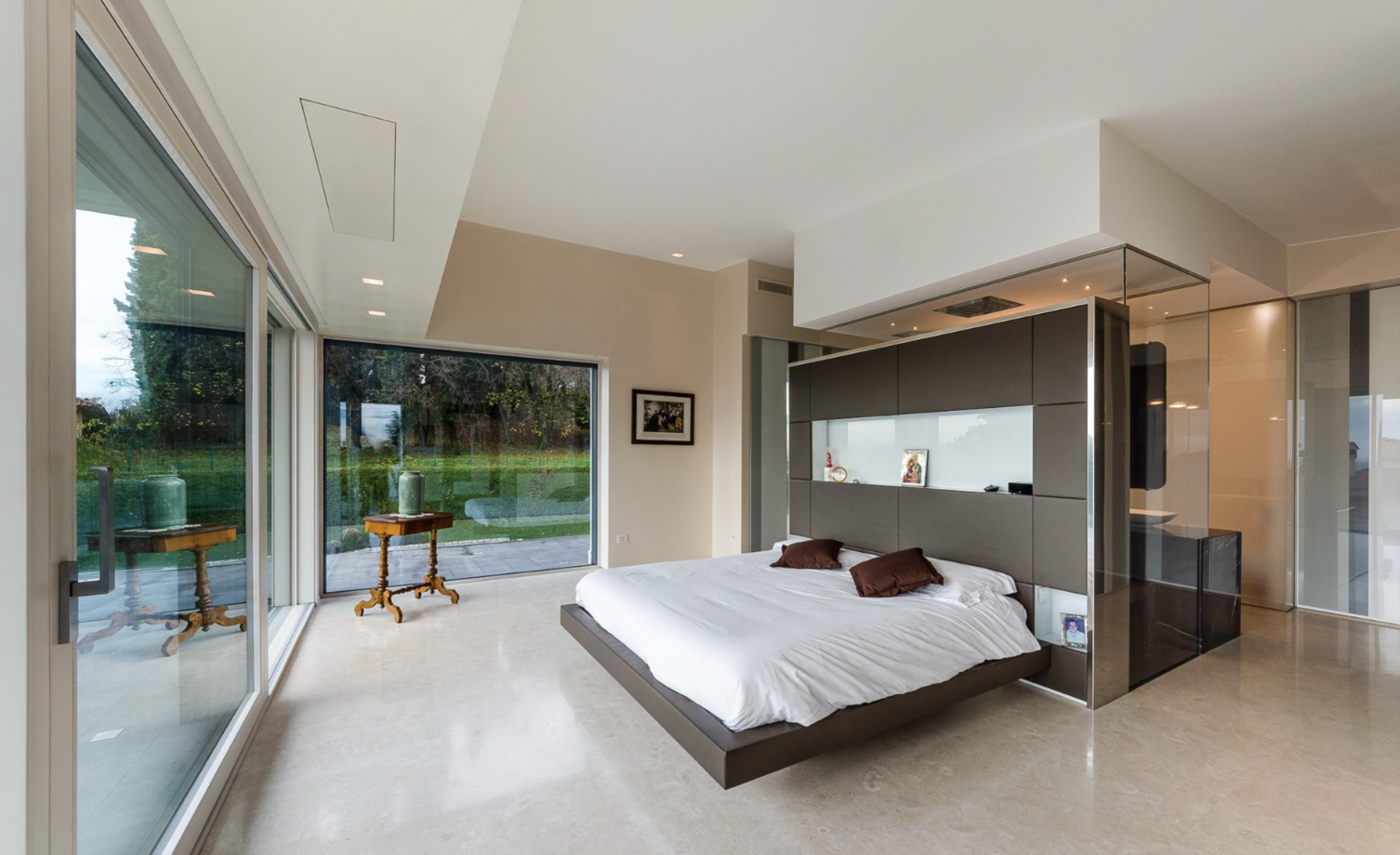Modern bedroom with a large glass sliding door opening to a green outdoor space, a bed with two brown pillows, and a gray partition wall with decorative shelves and built-in lighting.