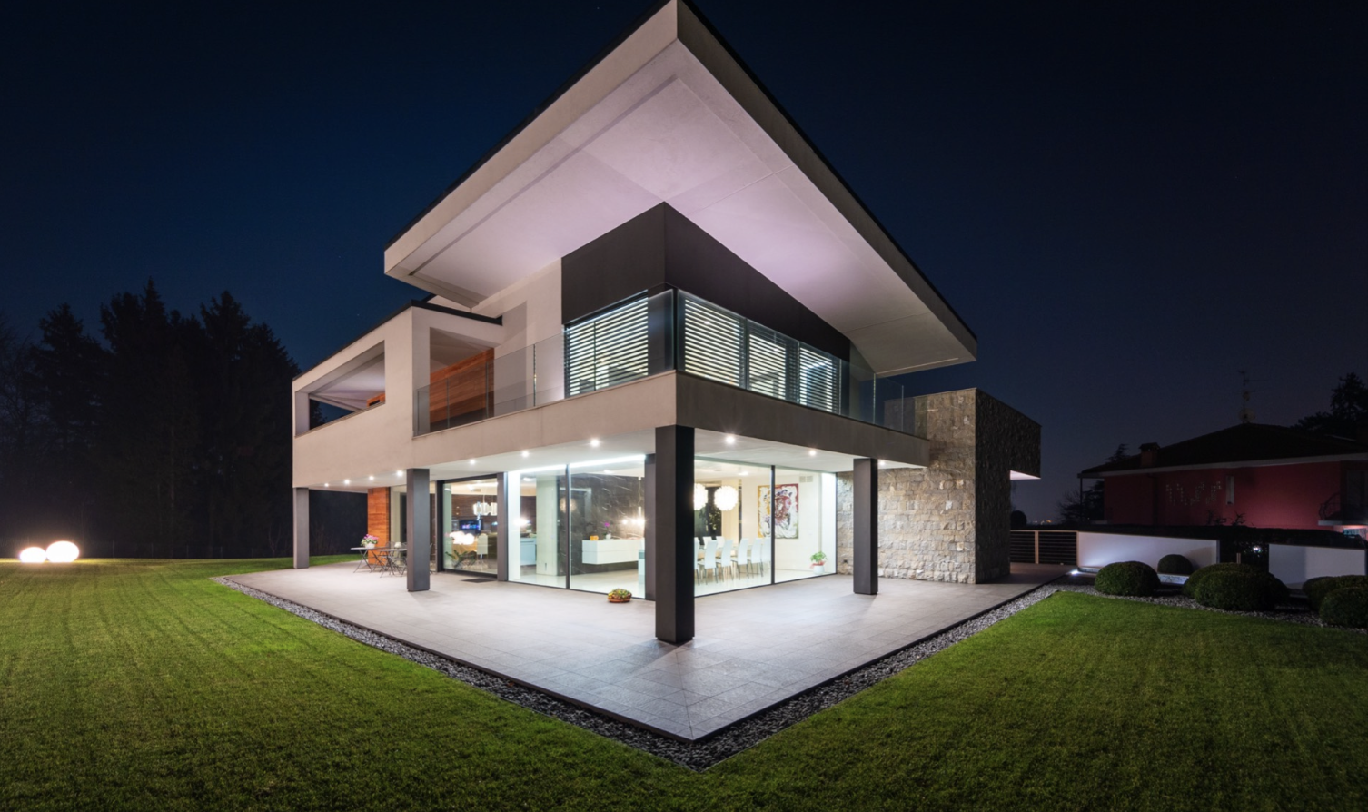 Night view of a modern two-story house with large glass windows, white exterior, and outdoor patio, surrounded by a well-maintained grassy lawn and trees in the background.