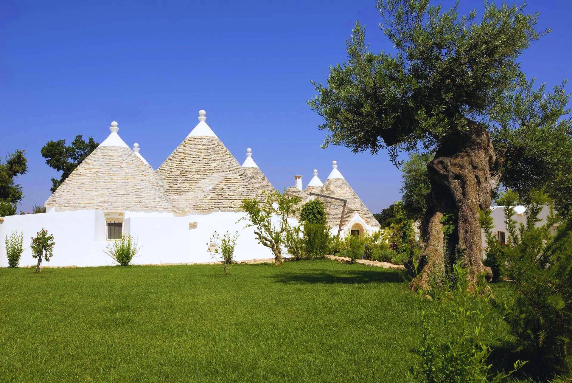 A traditional white stone building with conical roofs, set against a bright blue sky, surrounded by green grass, small trees, and an old gnarled tree in the foreground.