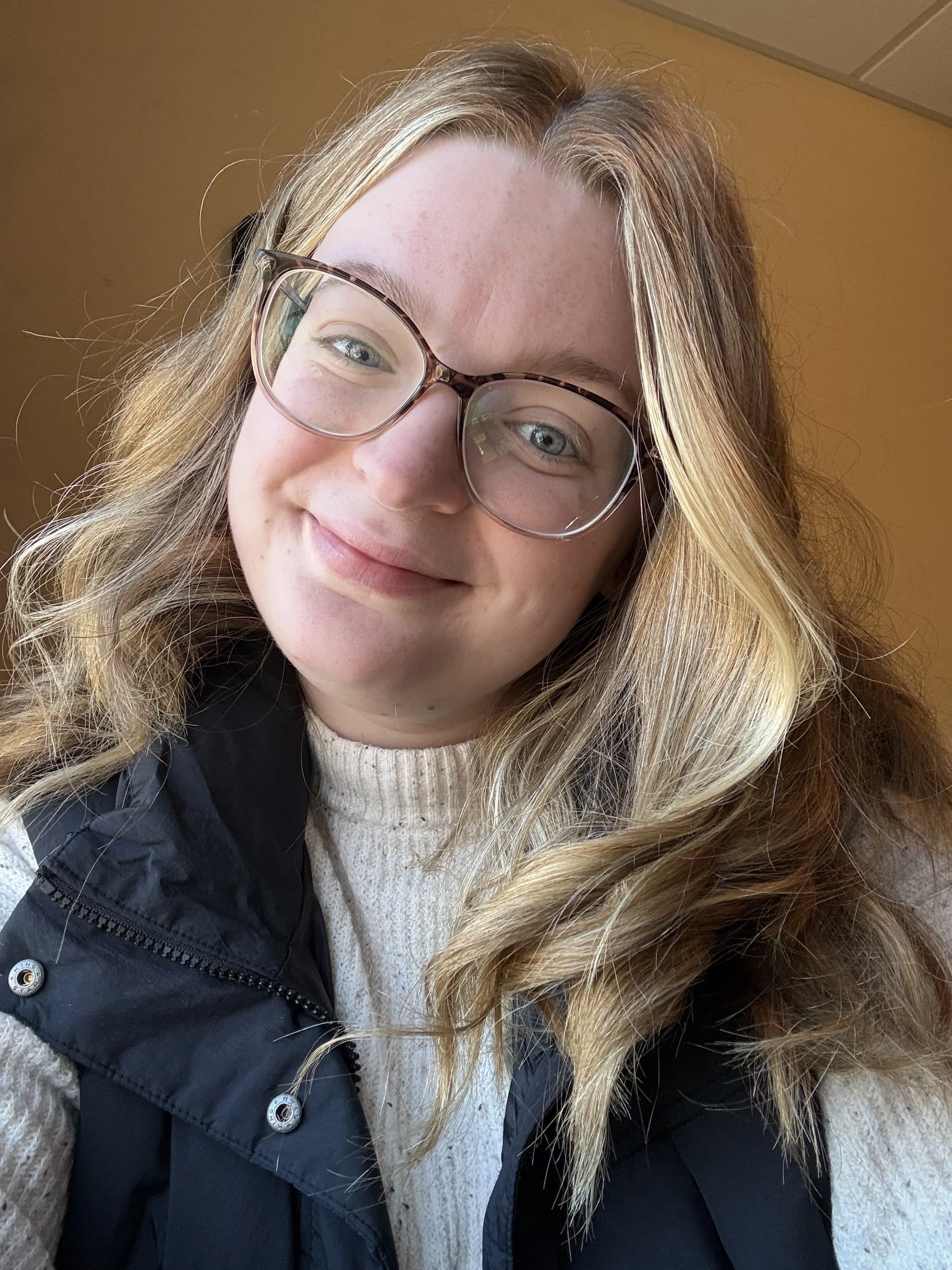 Friendly therapist with long blonde hair and glasses smiling in a softly lit indoor setting, wearing a cream sweater and black vest, conveying a warm and approachable demeanor.