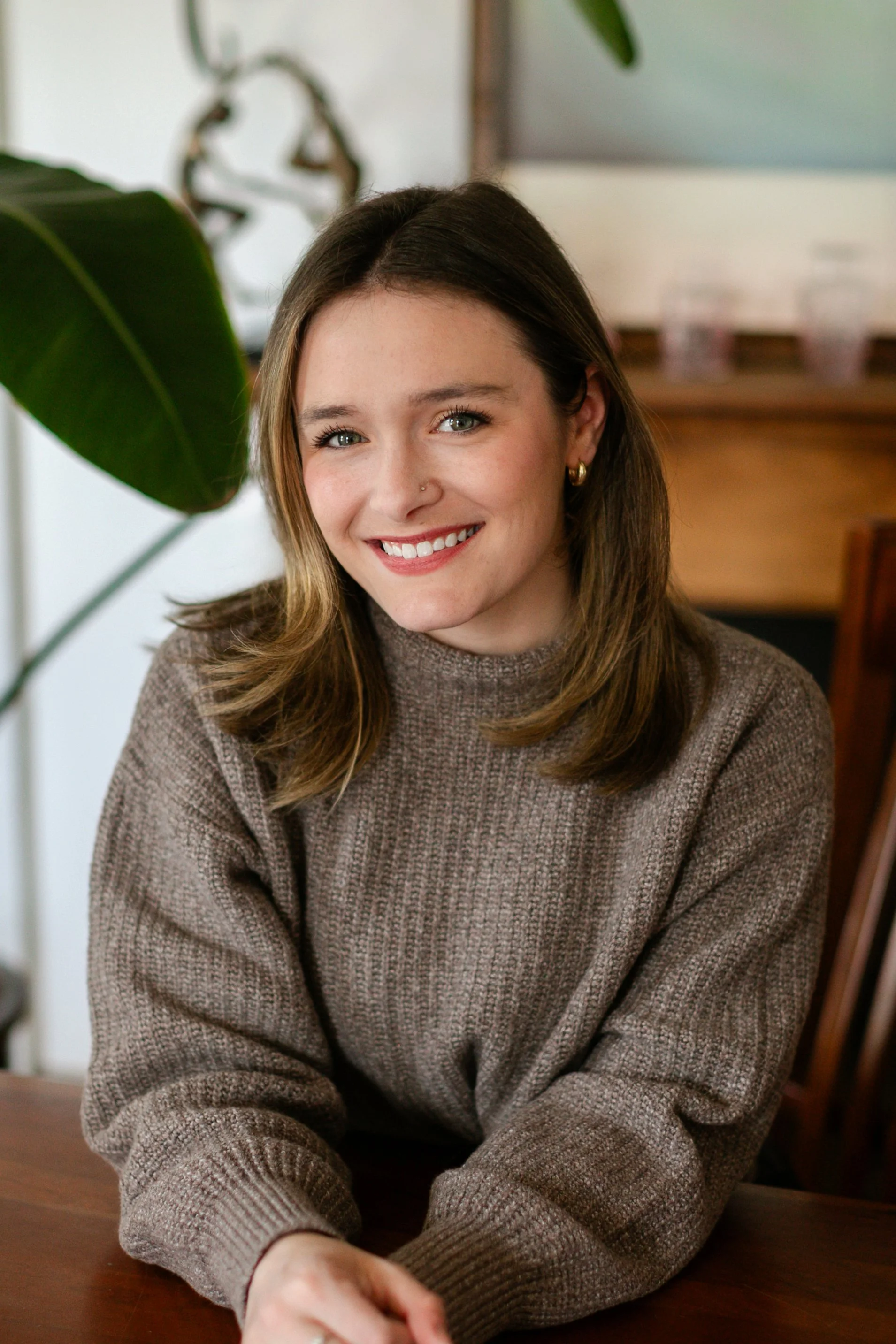 Smiling therapist with shoulder-length brown hair wearing a cozy knit sweater, seated at a table in a warmly lit space with greenery in the background, conveying an approachable and supportive presence.