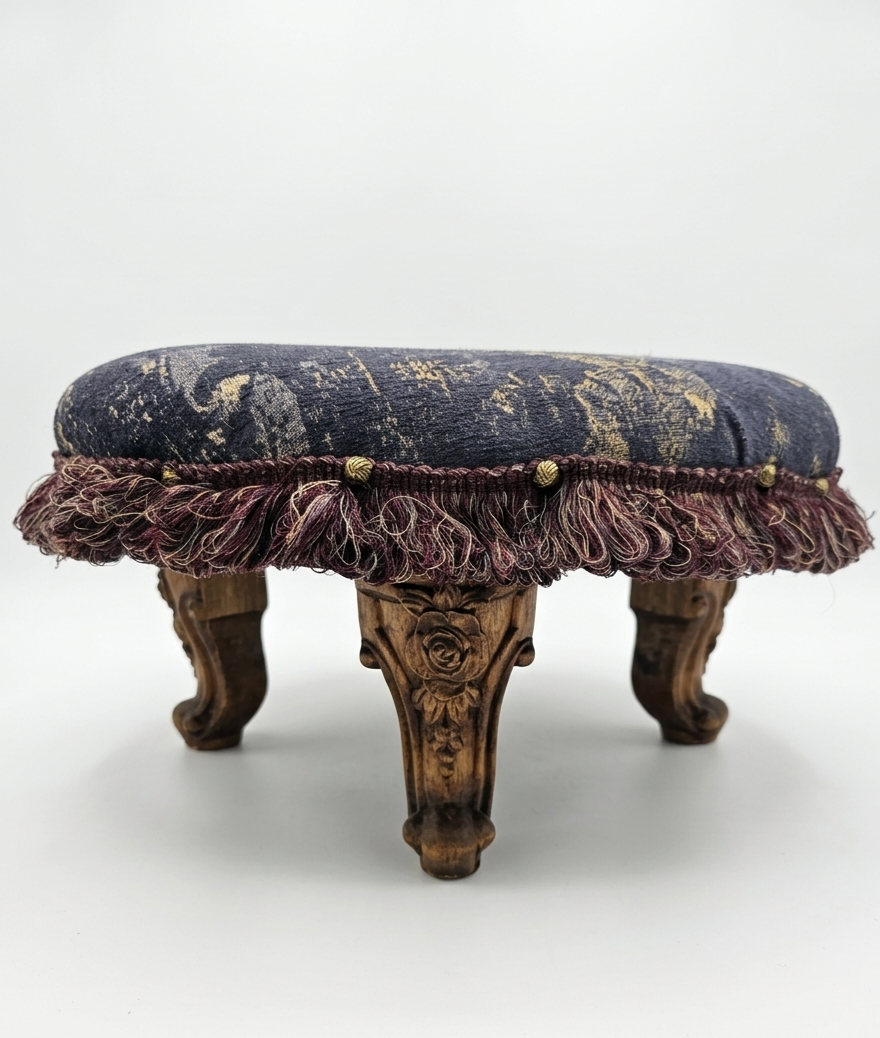 A small antique wooden stool/ottoman with ornate carved legs and a velvet cushioned top, edged with decorative fringe.