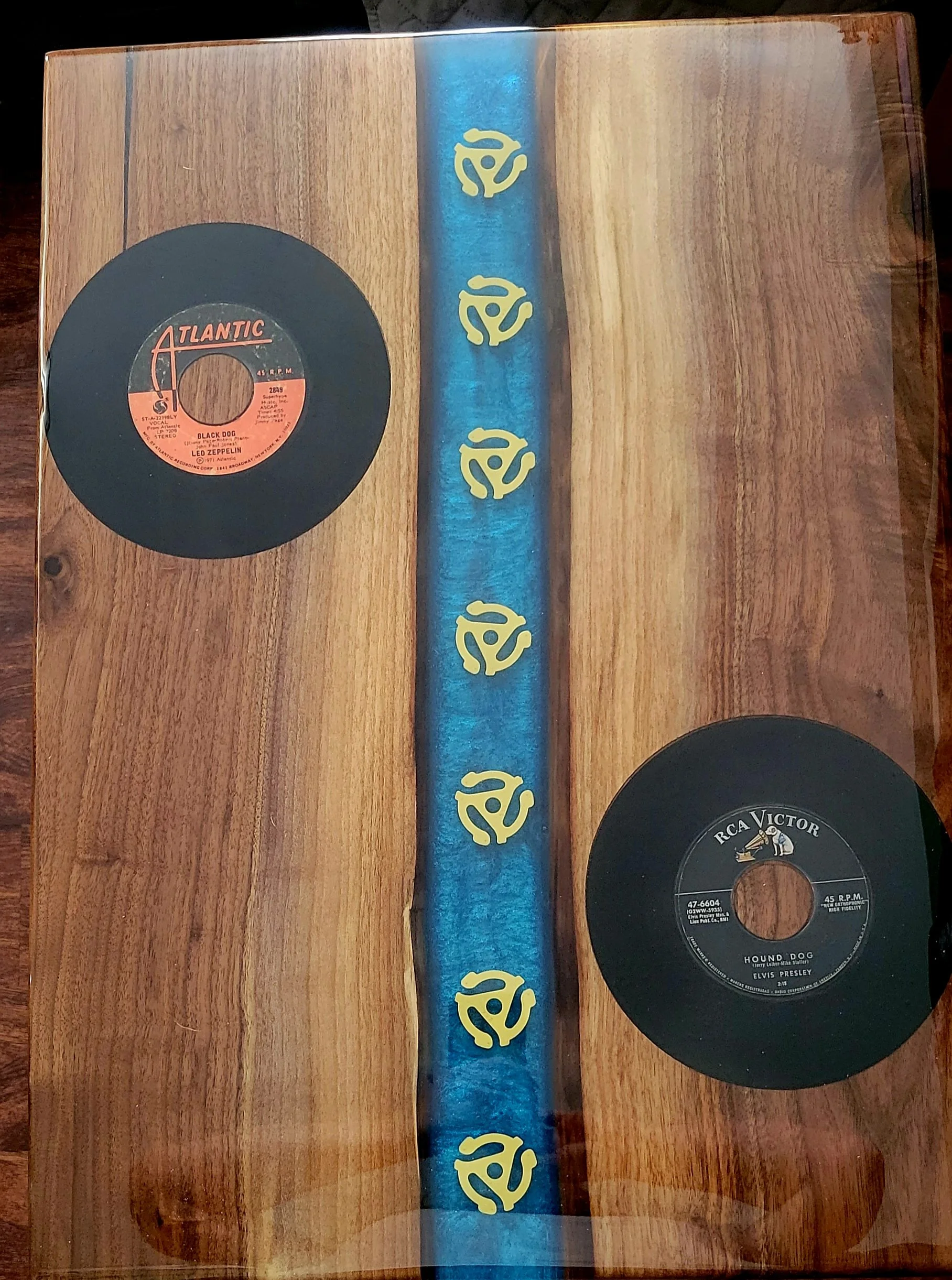A wooden table with two vinyl records, one labeled 'Atlantic' featuring Led Zeppelin's 'Black Dog' and the other labeled 'RCA Victor' featuring Elvis Presley's 'Hound Dog'. A blue cloth with yellow decorative symbols runs vertically between the records.
