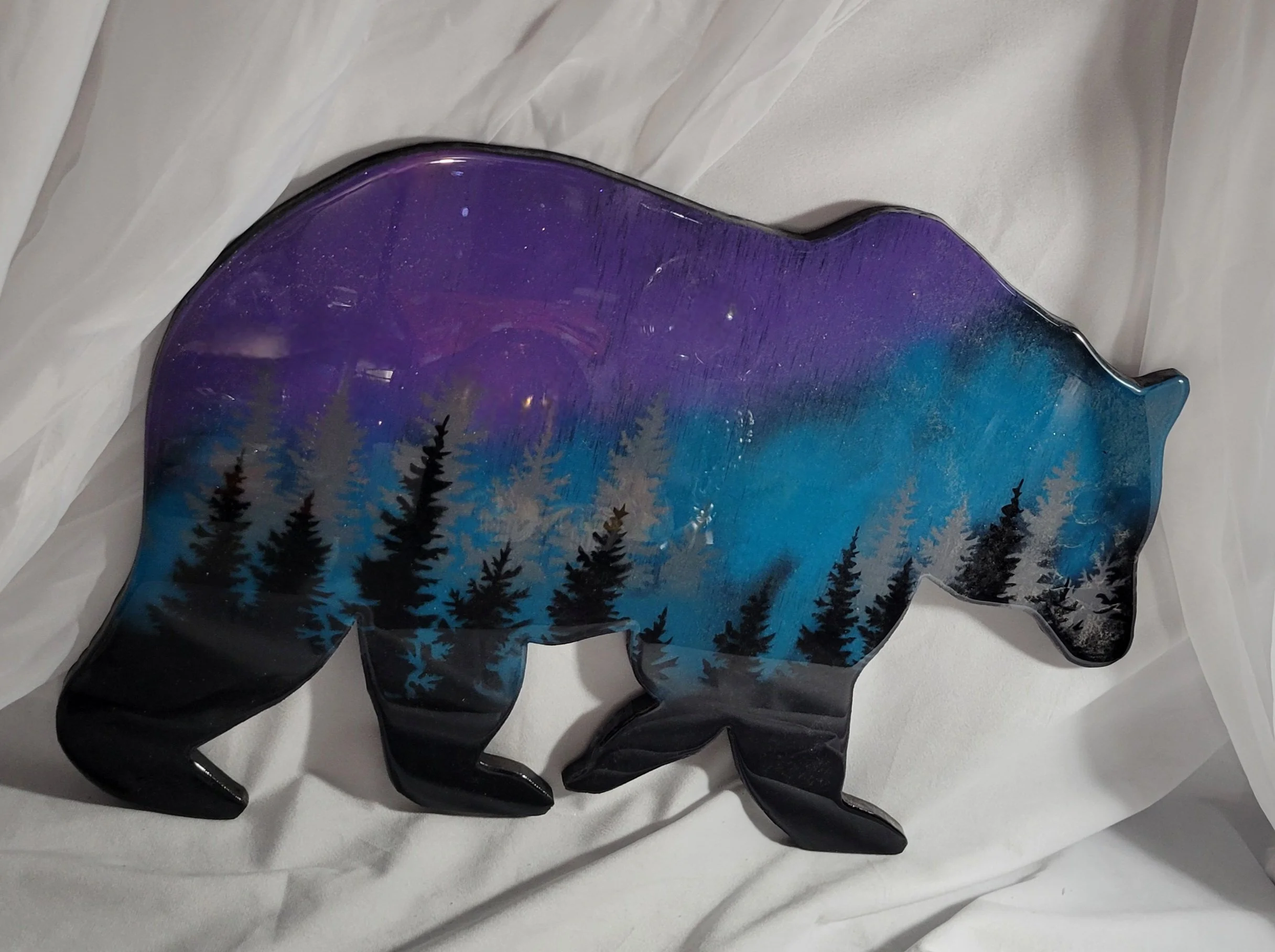 Painted bear-shaped object with a mountain and pine tree landscape in shades of purple, blue, and black.