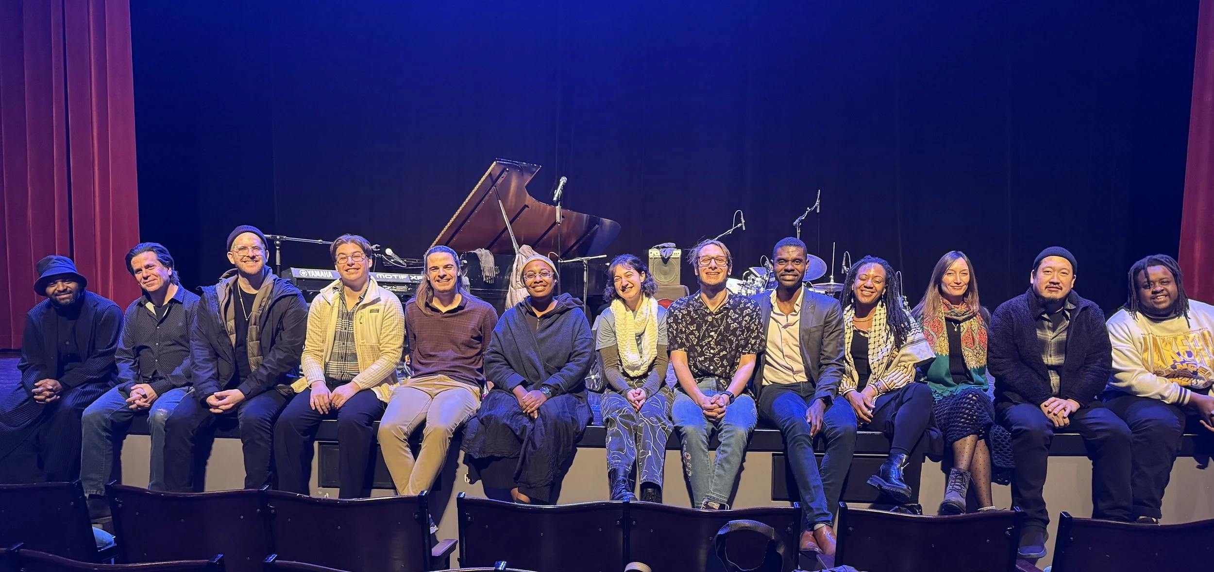 A group of twelve people seated in a row on the edge of a stage in front of a grand piano and drum set. The stage has red curtains on both sides and is lit with blue overhead lighting.