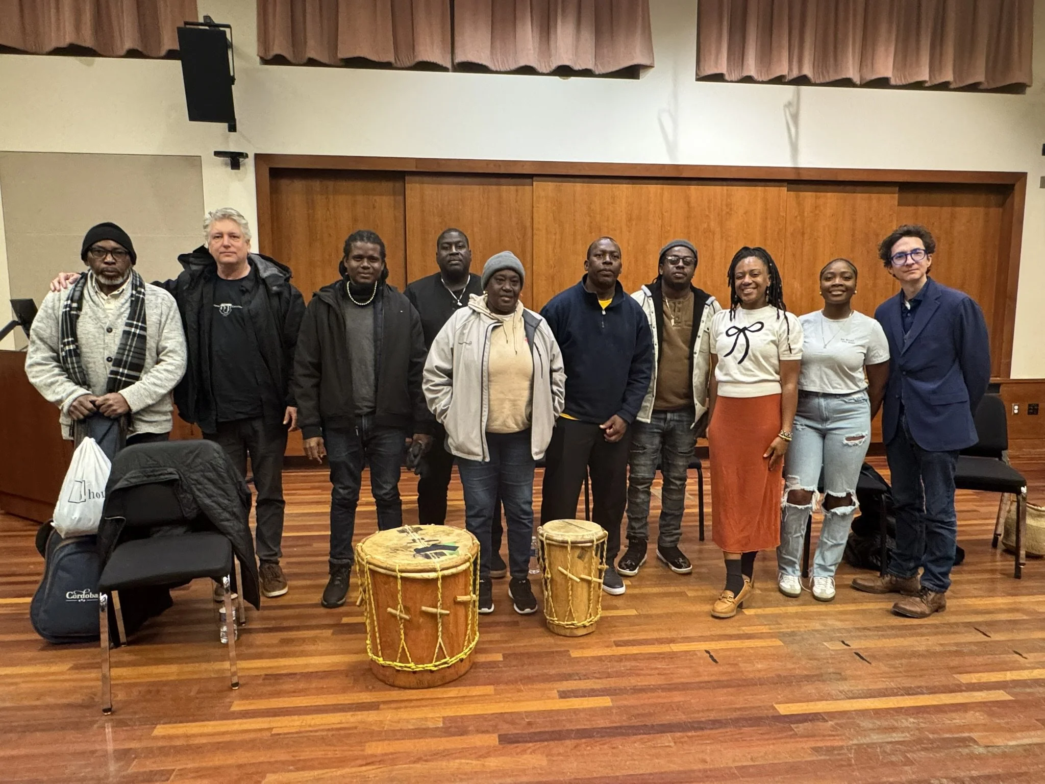 A group of people stands together in a music rehearsal room with wood floors and wood-paneled walls. Two traditional hand drums are placed on the floor in front of the group