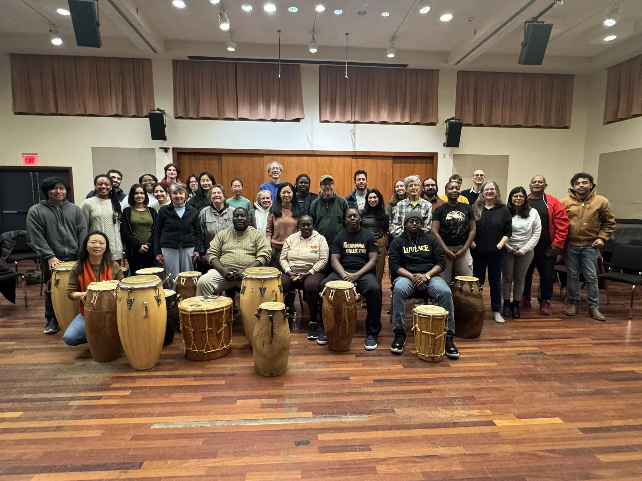 A large group of people poses together in a music rehearsal room, with several hand drums of various sizes arranged in front of the group. The room has wooden floors, acoustic panels, and overhead lighting, and the group appears gathered after a drum