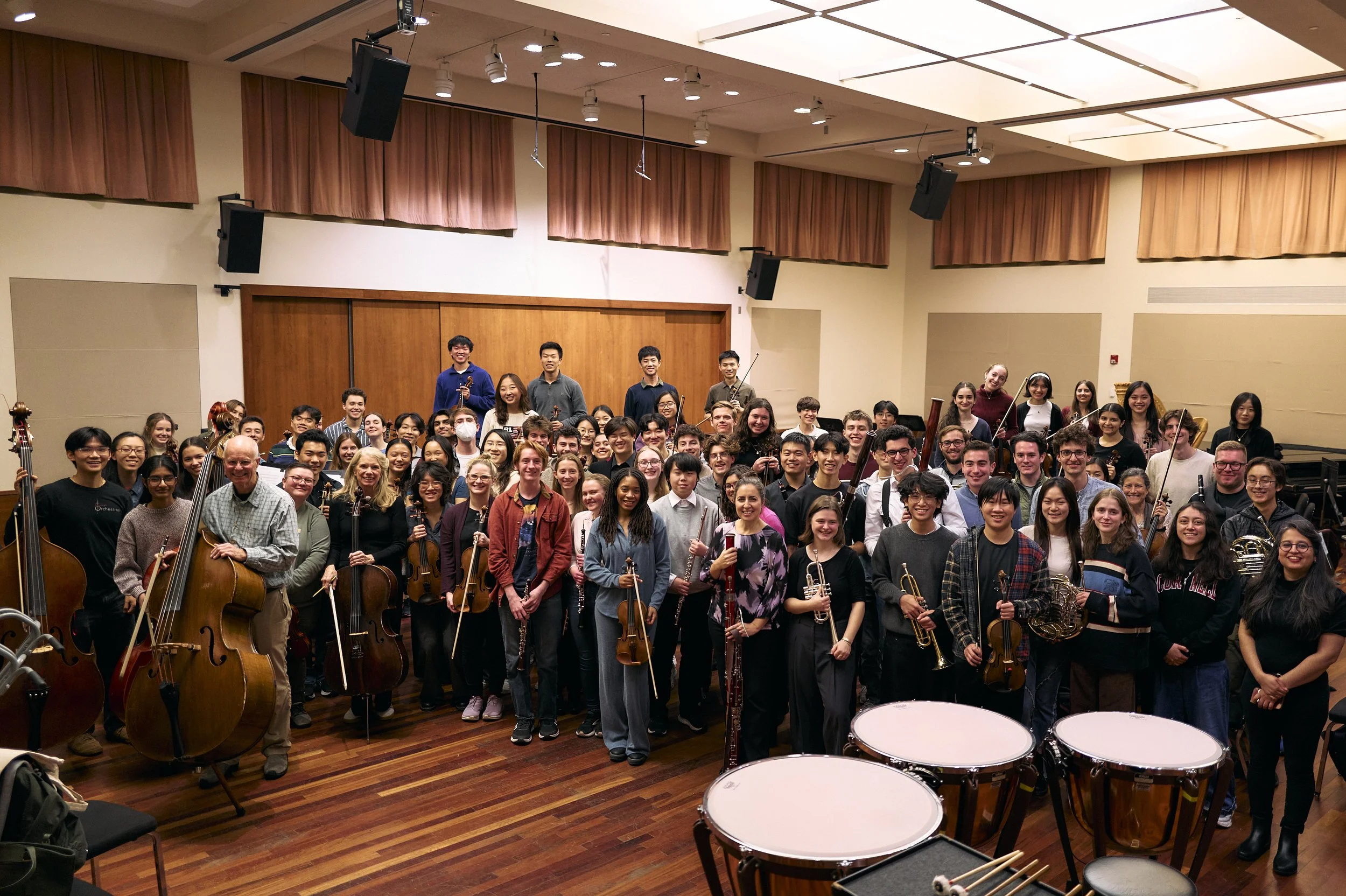A large group of musicians gathered in a rehearsal room, holding various instruments including violins, cellos, double basses, woodwinds, and brass. Timpani drums are visible in the foreground.
