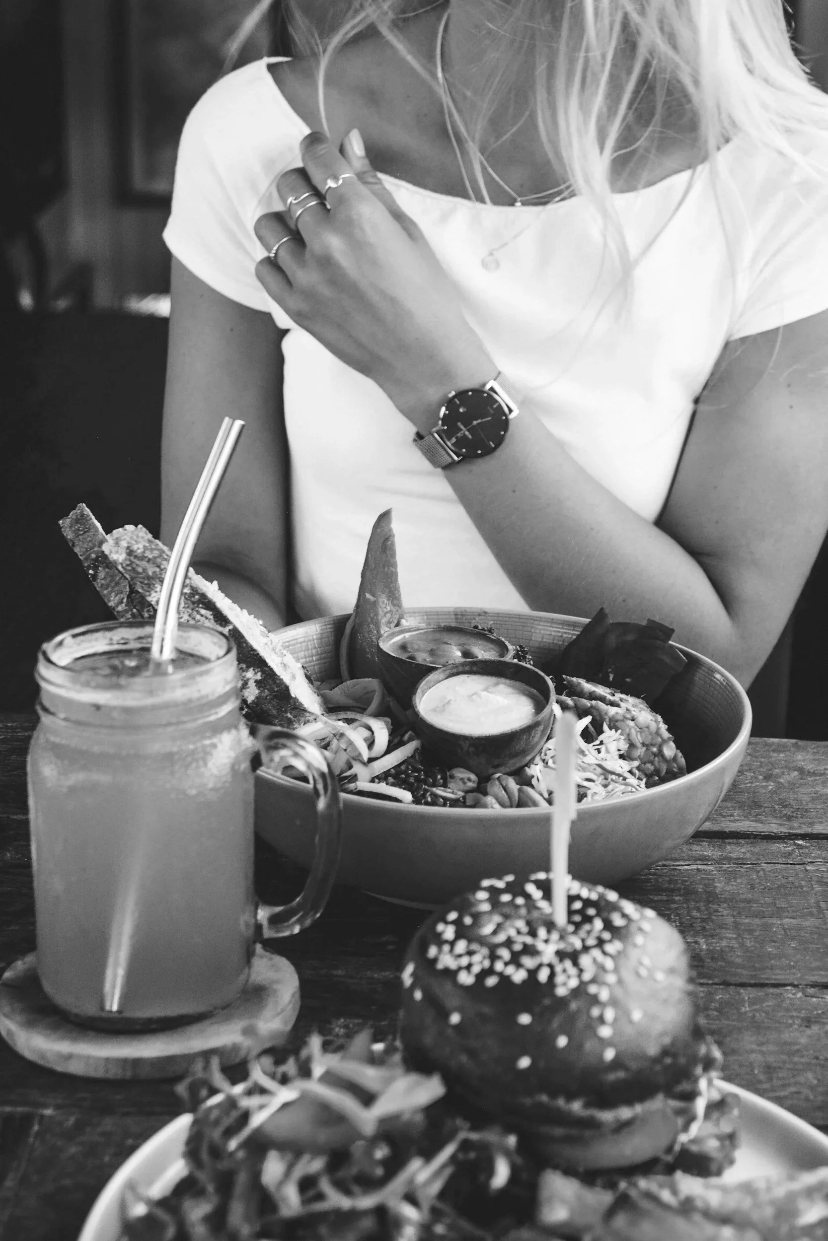 Black and white photo of a person with long hair wearing a watch and rings, sitting at a table with a large bowl of assorted food, a jar of what appears to be a beverage, and a burger with sesame bun and toppings.
