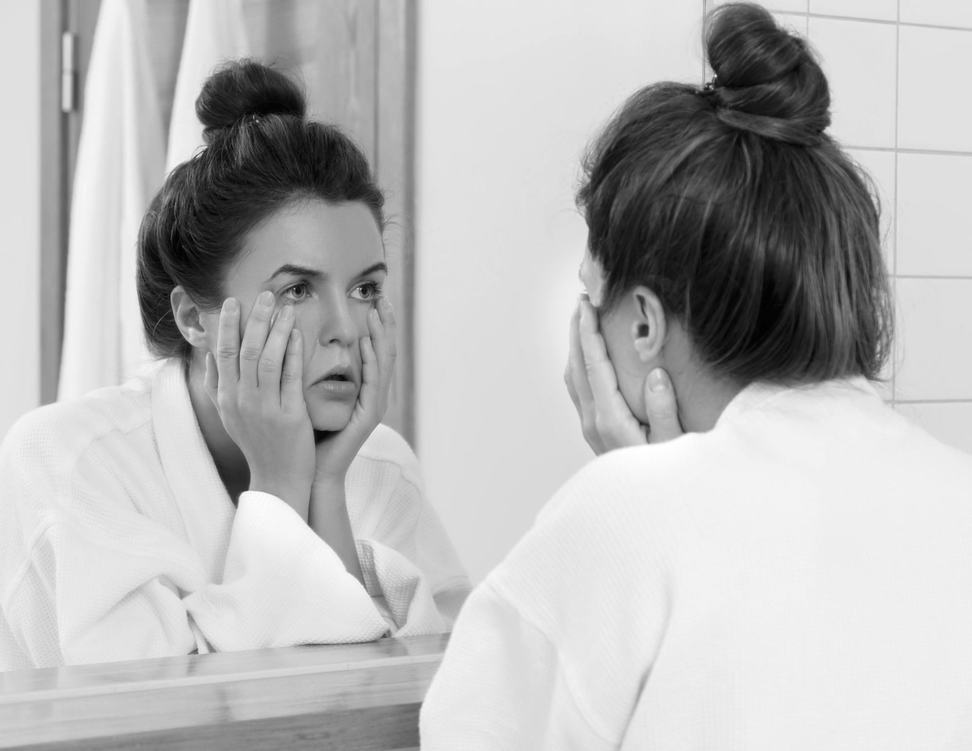 A woman with dark hair in a bun, wearing a white outfit, looking at herself in a mirror and touching her face with both hands. She appears to be examining her face closely.