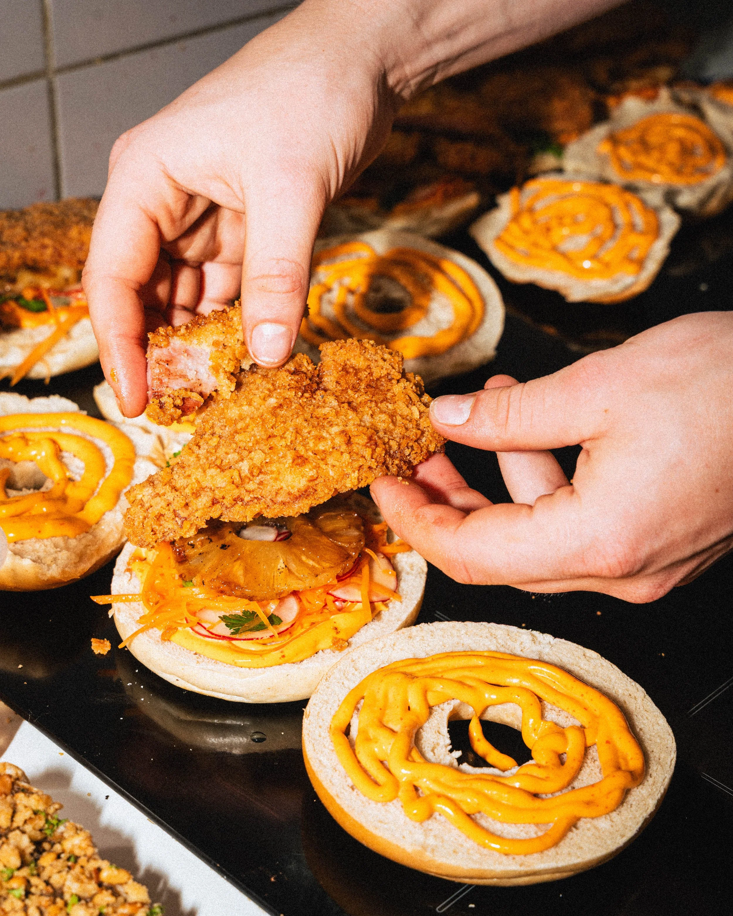 Person assembling an open-faced sandwich with fried chicken, pineapple, shredded vegetables, and mustard on a bagel