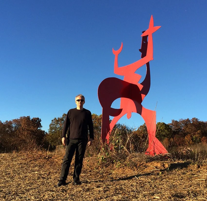 Man standing next to a large, abstract, red sculpture of a reclining woman in an open field with trees in the background and a clear blue sky.