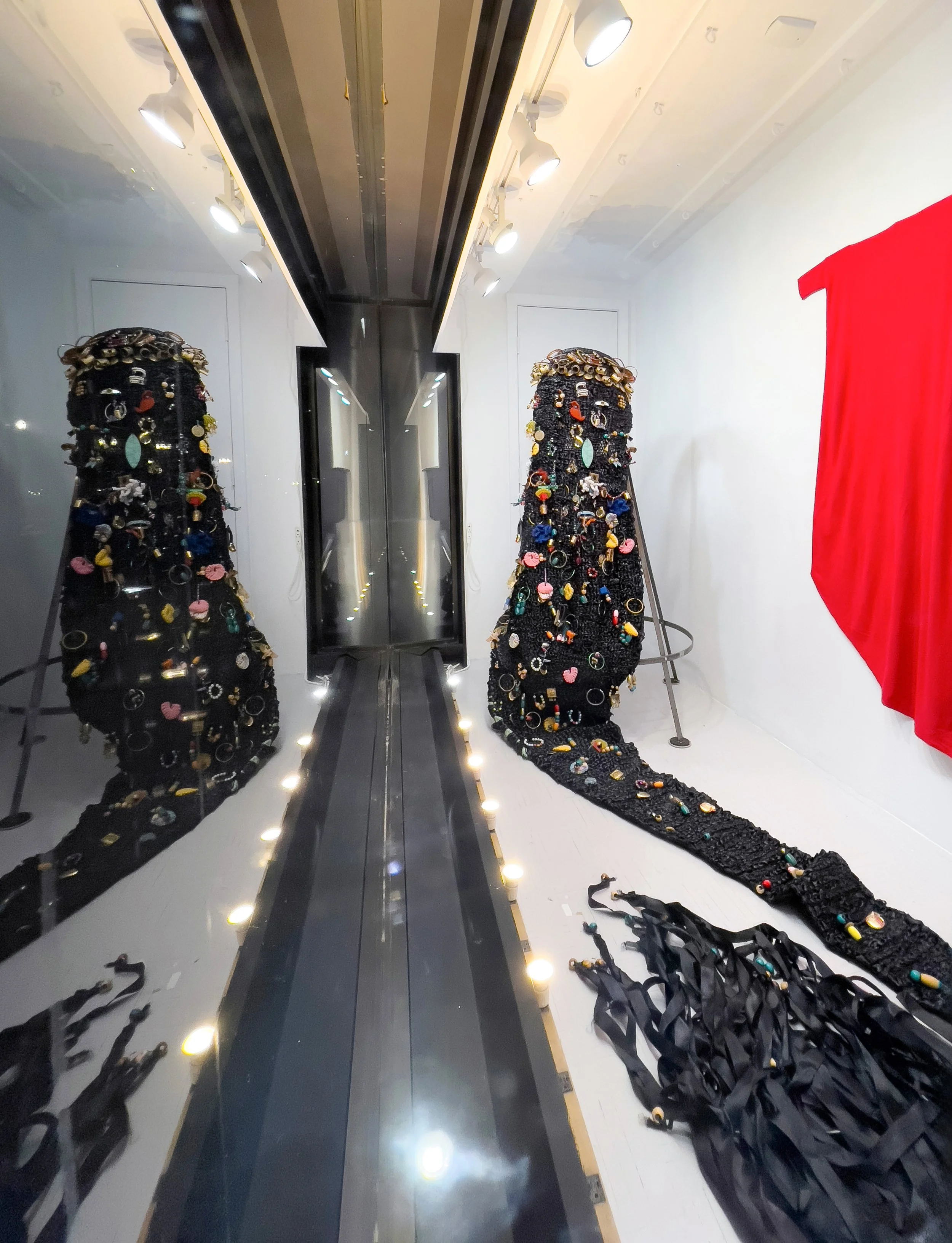 Installation art featuring a long, black, textured sculpture resembling a snake, decorated with colorful beads and accessories, flanked by mirrors and illuminated by small lights, with a red backdrop on the right.