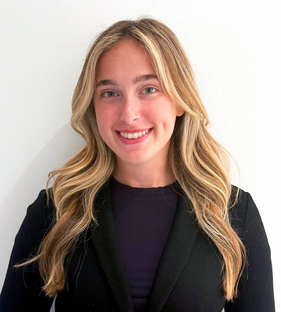 Young woman with long blonde wavy hair, smiling, wearing a black jacket and black shirt, standing against a white wall.