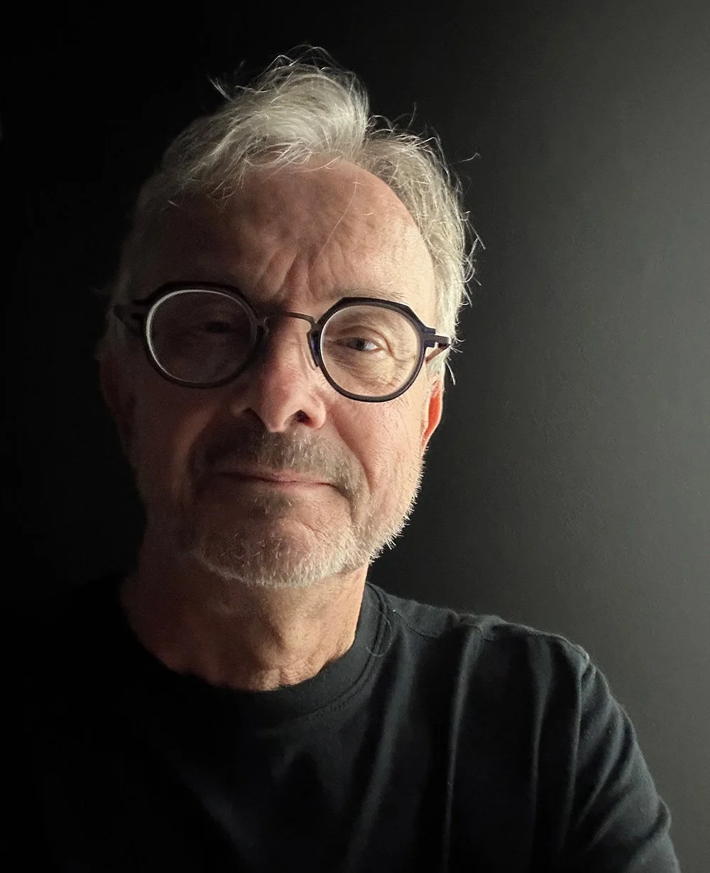 Close-up of a middle-aged man with white hair, beard, round glasses, wearing a black shirt, against a dark background.