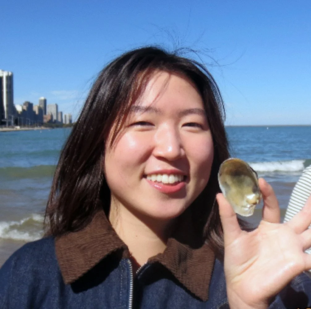 Smiling woman on the beach holding a clam shell, with city skyline and ocean in the background.