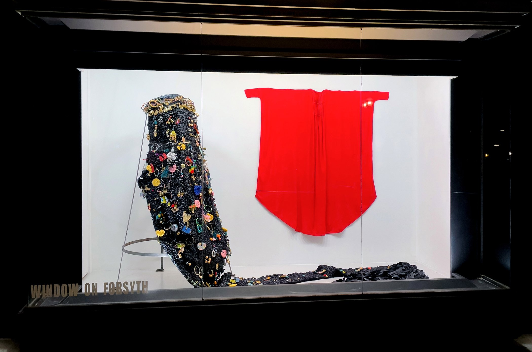 Display of two fashion pieces: a black garment adorned with colorful buttons and embellishments on the left, and a bright red fabric piece on the right, both behind glass at a museum exhibit.