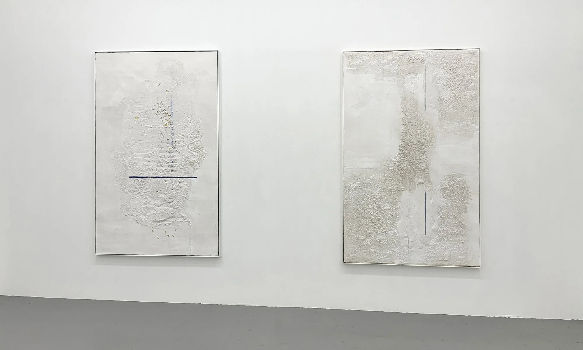 Two minimalist abstract paintings on a white gallery wall, featuring textured off-white surfaces with subtle hints of color and blue lines.