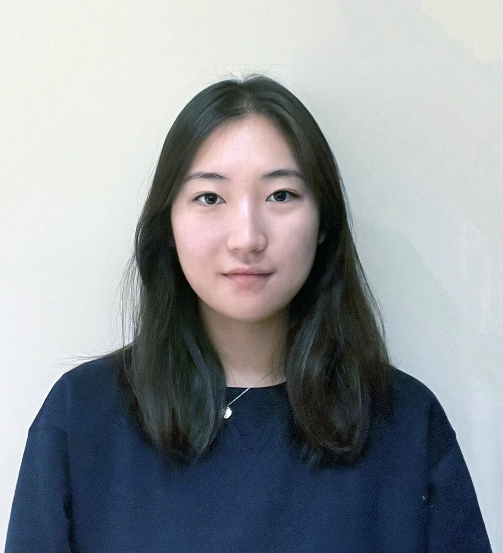 A young woman with straight, shoulder-length black hair wearing a dark sweatshirt and a delicate necklace, standing against a plain off-white background.