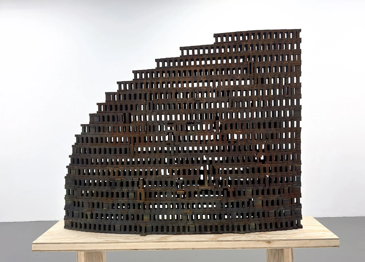 Contemporary sculpture with irregular, stacked layers made of dark metal, placed on a light wooden stand against a plain white background.