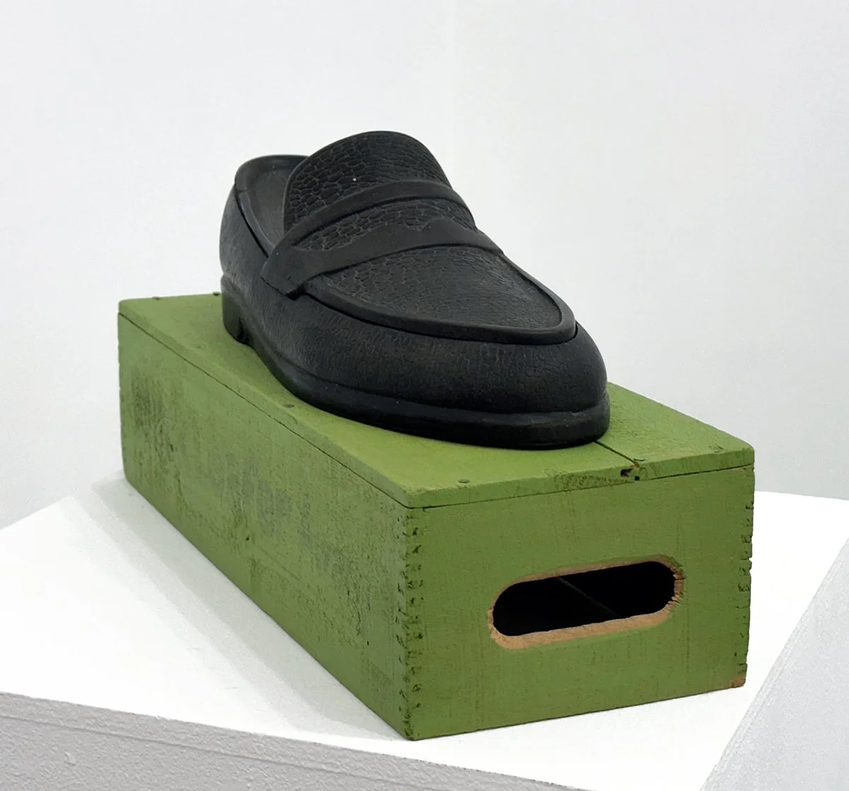 sculpture of Black loafer shoe displayed on a green wooden box, against a white background.
