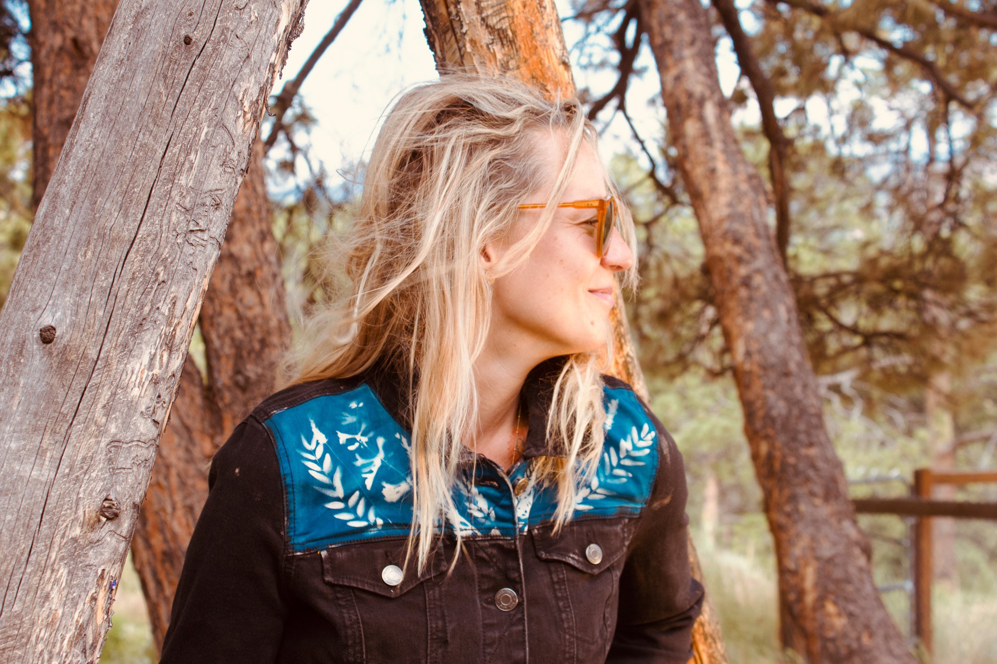 Woman in glasses and denim jacket standing by trees outdoors.