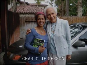 Two women smiling and standing outside, one older woman with gray hair and a younger woman holding a book. They are near a black car and a wooden fence.