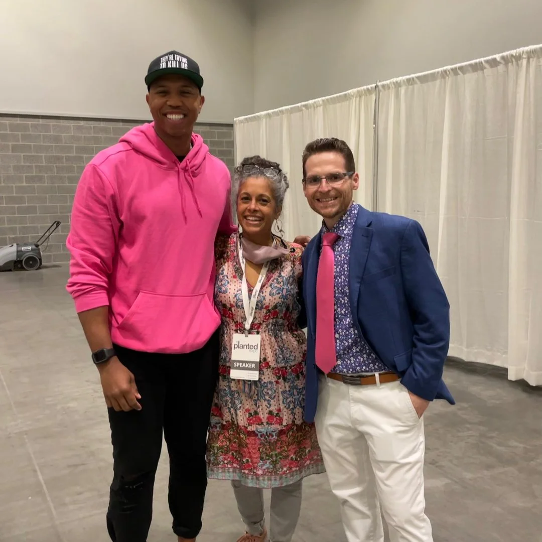 Three people standing together indoors, smiling at the camera. One man is wearing a bright pink hoodie and a hat that says 'They're Trying To Kill Us.' A woman in the middle has gray hair, is wearing a floral dress with a conference badge that says 'SPARK' and 'SPEAKER.' Another man on the right is dressed in a blue blazer, floral shirt, pink tie, and white pants. There is a curtain behind them.