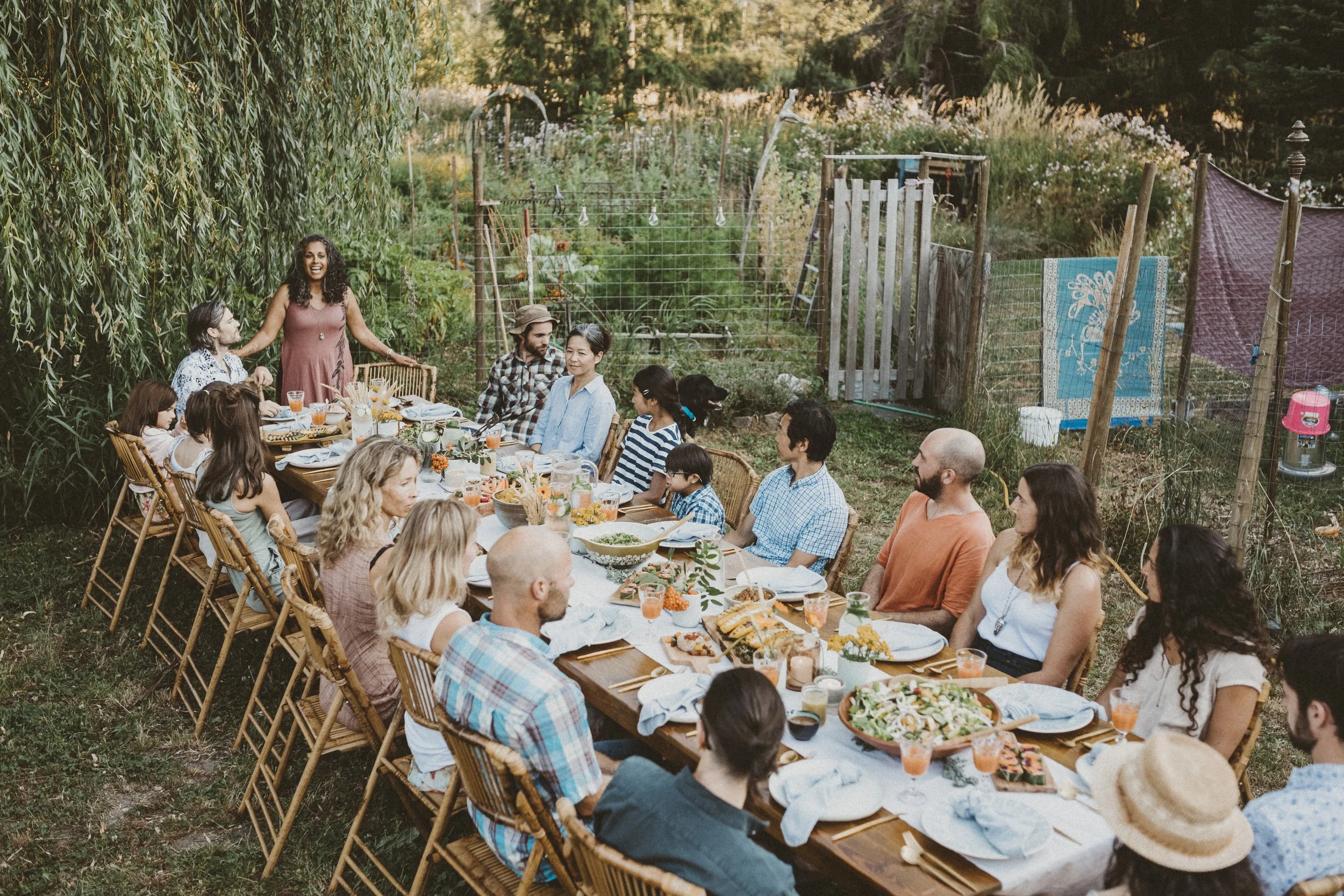 A large outdoor gathering with around 20 people seated at a long table in a lush garden. The table is set with dishes, drinks, and flowers. Women and children are on one side, and men on the other. One woman in a pink dress stands and speaks, smiling to the group. Behind, there are trees, plants, and garden fencing.