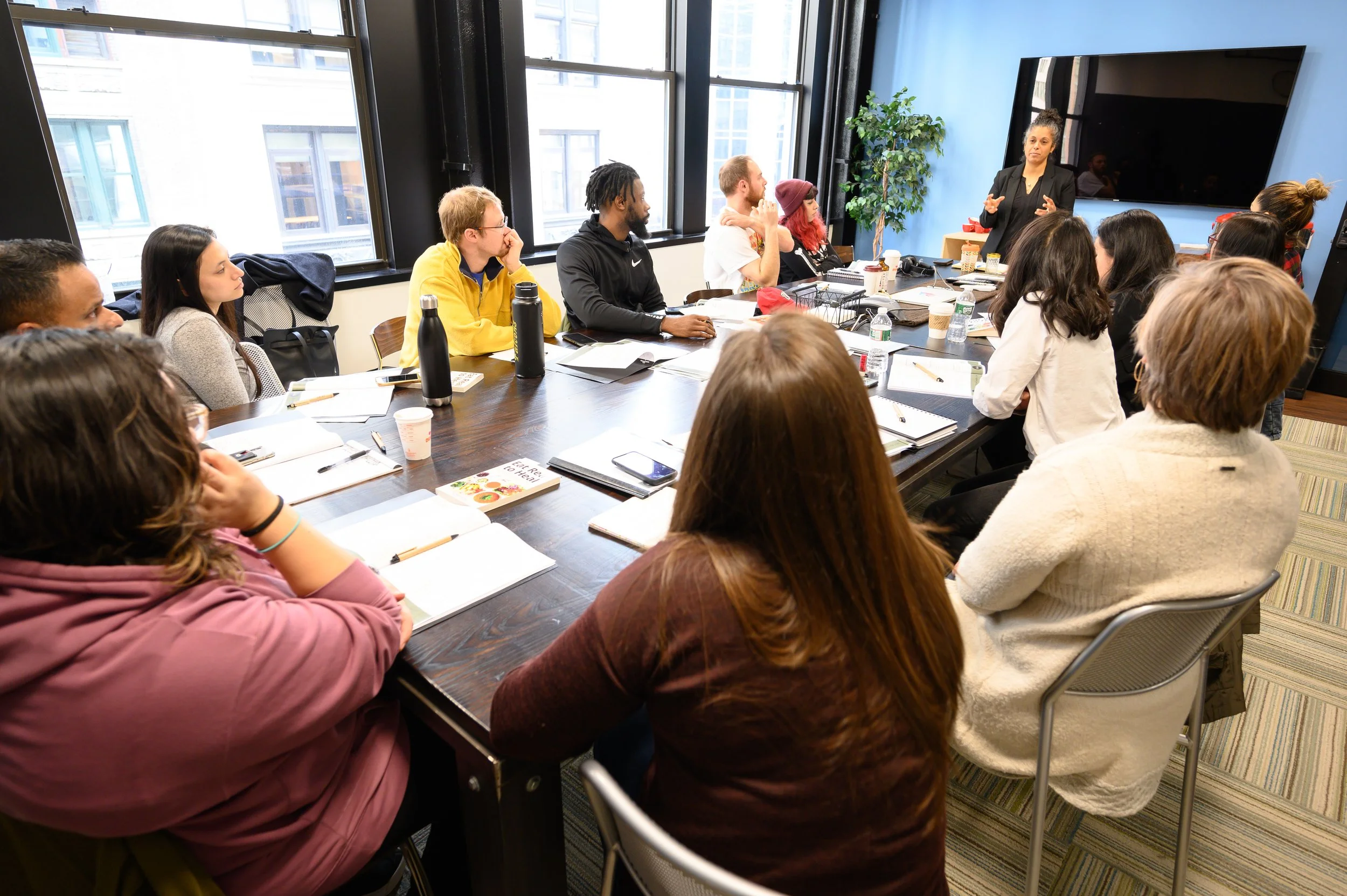 A diverse group of people attending a meeting or workshop in a modern conference room, listening to a woman standing and speaking at the front.