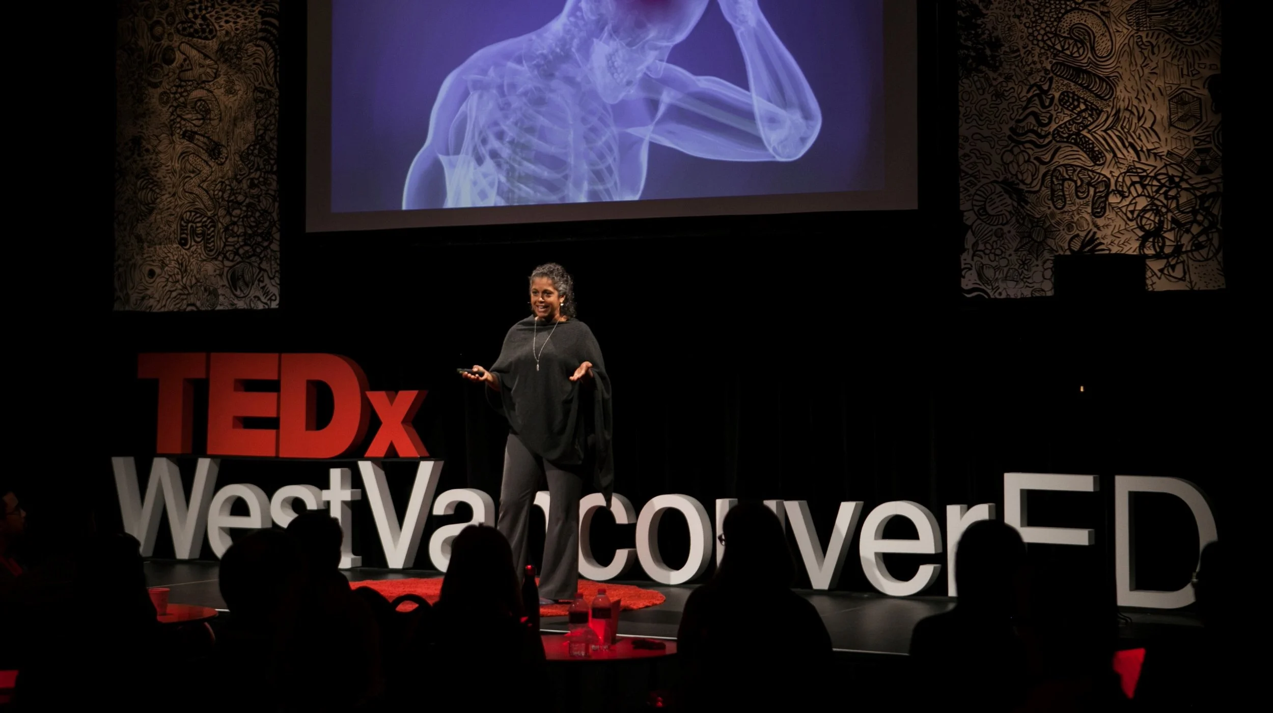 A woman giving a TEDx talk on stage with large red and white TEDx letters and the words 'West Valley Conver' behind her, a large screen showing a medical illustration of a person's upper body, and a dark background with intricate artwork on the sides.