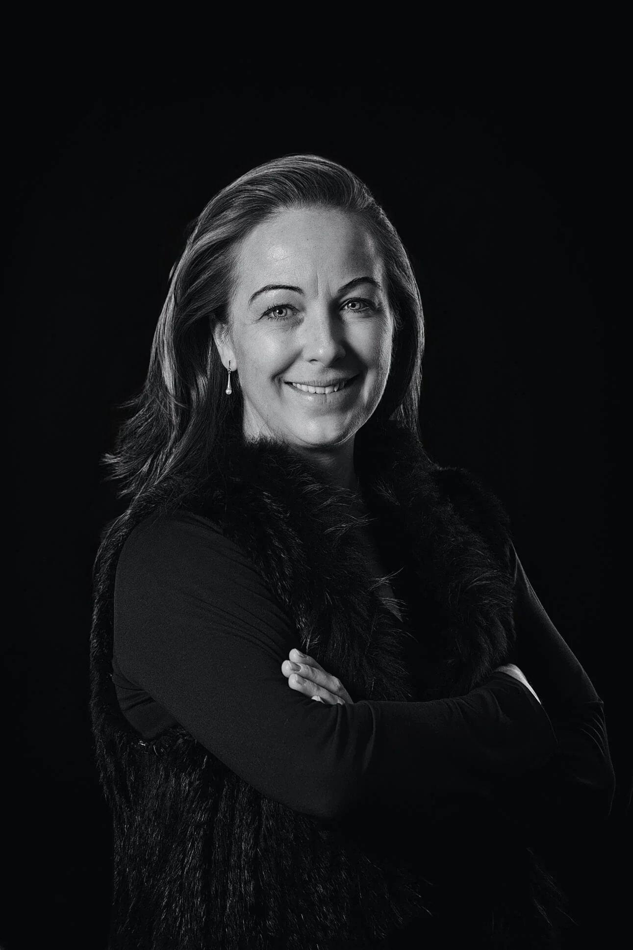 Portrait of a woman with shoulder-length hair smiling, wearing earrings and a dark outfit with a faux fur collar, black background.