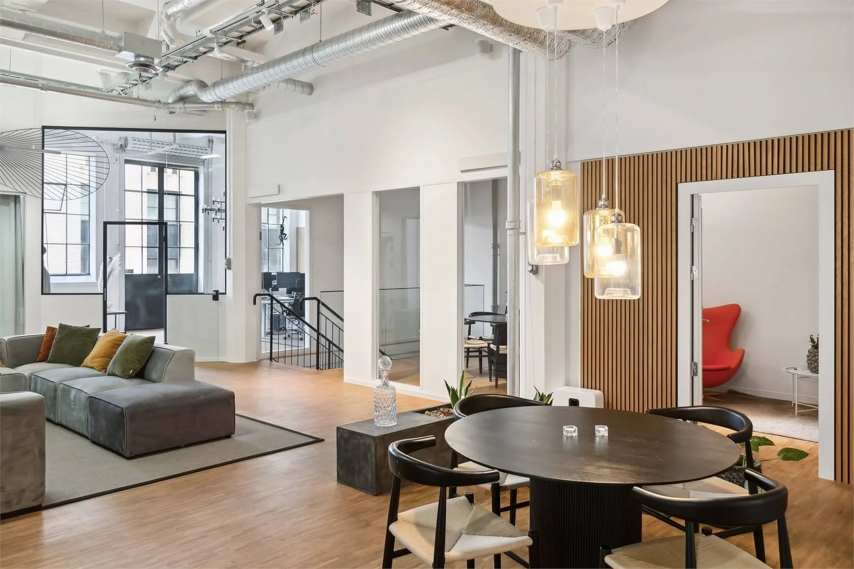 Modern co-working space with a round dining table, black chairs, a beige sofa with colorful pillows, pendant lights, and a small room with a red chair and pineapple decor.