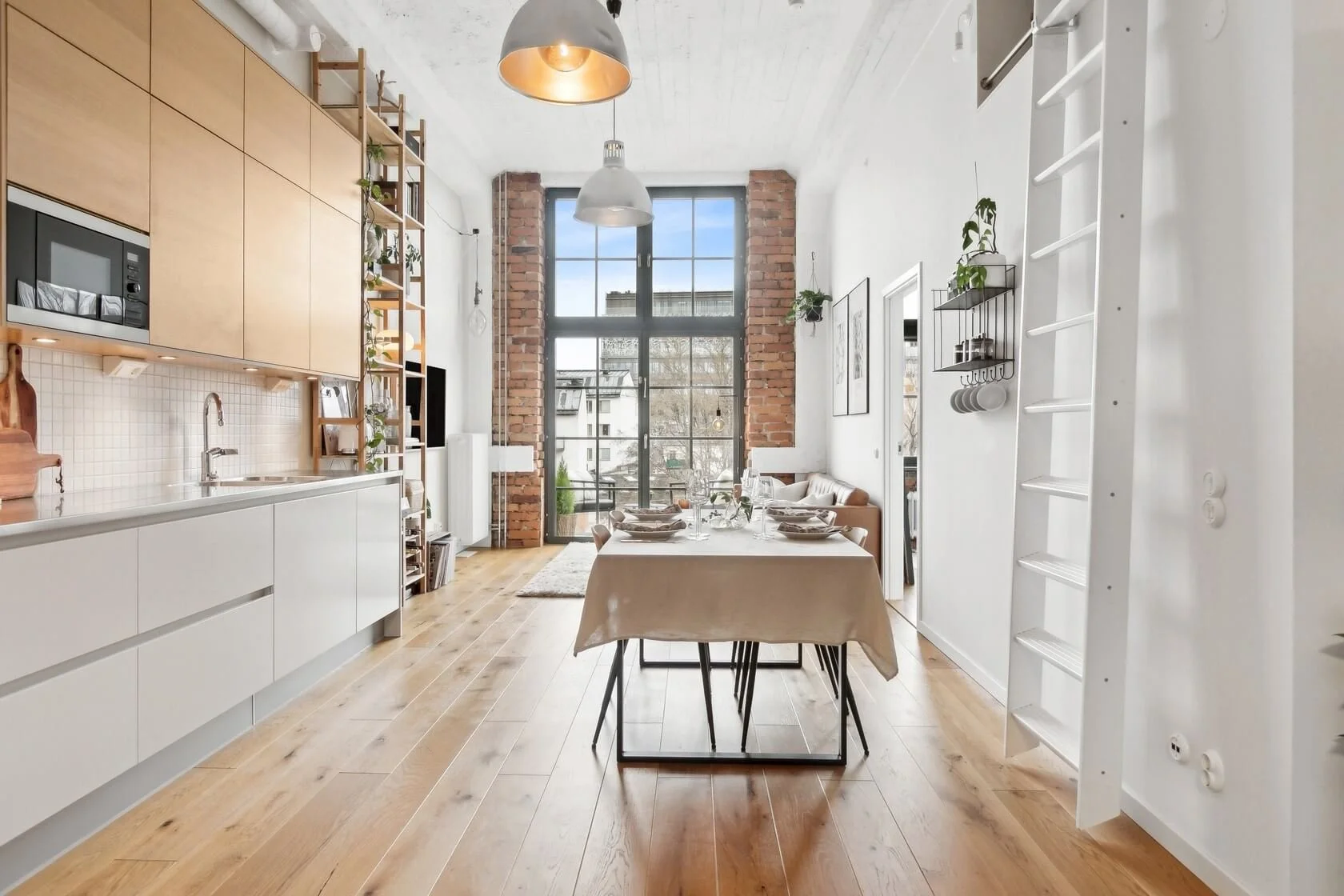 Modern dining area in an urban apartment with large window, wooden floors, and exposed brick wall, featuring a table set for meals.