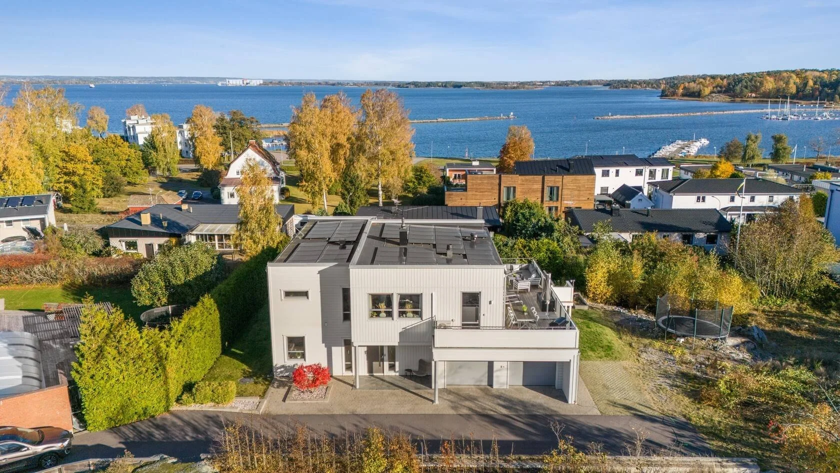 Aerial view of a modern white house with a flat roof and rooftop terrace situated near a lakeshore, with other houses, trees, and boats visible in the background on a clear day.