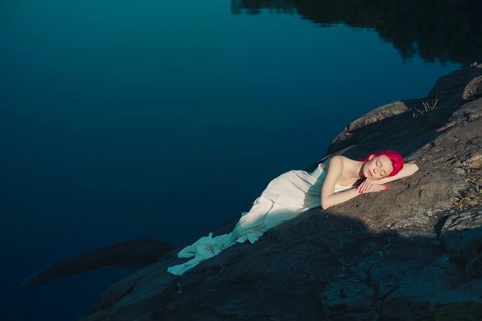 A woman with bright pink hair, wearing a white dress, is lying on a rocky slope near a calm body of water with her eyes closed, appearing to be sleeping or resting.