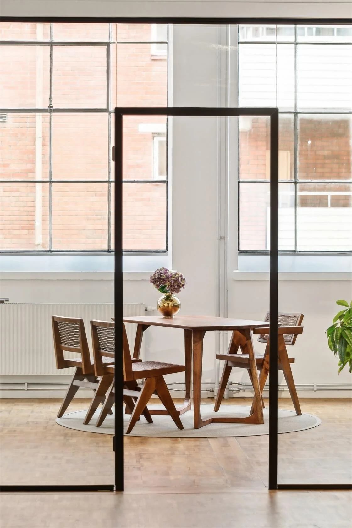 Interior view of a dining area seen through glass sliding doors, with a wooden dining table, four matching chairs, a vase with flowers on the table, a large window, radiator, and a potted plant.